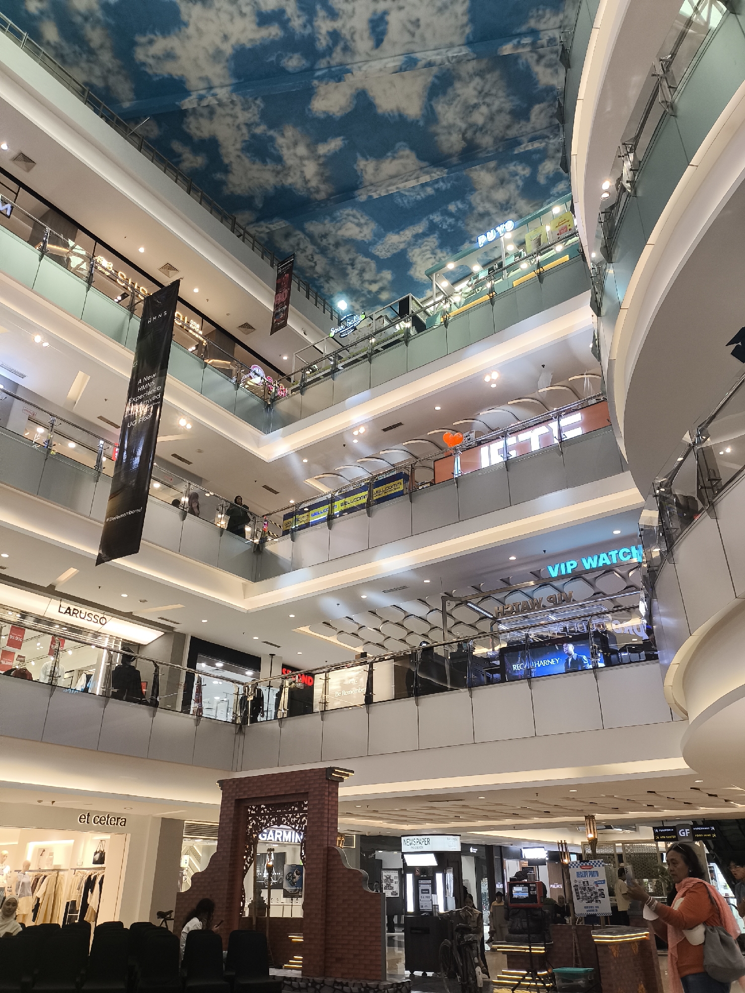 The image shows the interior of a multi-level shopping mall with a sky-themed ceiling. On the ground floor, a decorative brick archway stands in the foreground, with "GARMIN" signage visible on a display behind it. To the left of the archway, "et cetera" store signage is visible. Several black chairs are arranged in front of the archway, with a few individuals seated and standing nearby.
Further up in the mall, various shops are visible on different levels, indicated by signage such as "LARUSSO," "HMNNS," and "VIP WATCH." Banners hang from some levels, including one with "A New HMNS Experience Arrived." People can be seen browsing in shops, walking on the upper levels, and standing on balconies. The mall features modern architecture with curved white walls and glass railings. The overall lighting suggests it is daytime or the mall is well-lit. The context of Grogol, Indonesia, is not directly evident from the visual elements alone but is provided as the location.