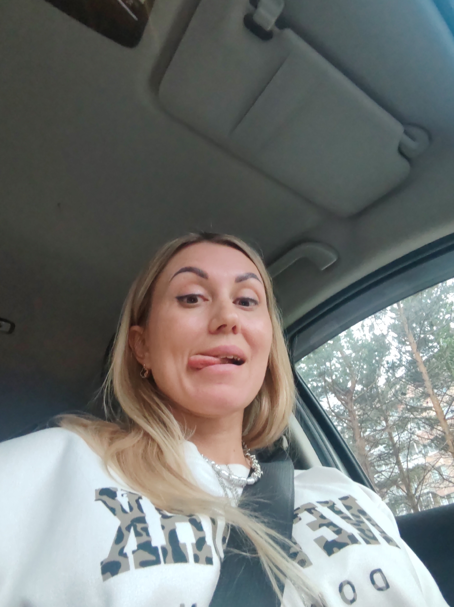 A woman with blonde hair is in the driver's seat of a car, looking at the camera with her tongue sticking out playfully. She is wearing a white sweatshirt with a camouflage-patterned design on it, along with a chunky silver necklace. Her seatbelt is fastened. Outside the car window, trees are visible, suggesting an outdoor setting. The interior of the car, including the sun visor and ceiling, are also visible. The lighting suggests it is daytime. The image has a casual, selfie-like feel. The visible text on her sweatshirt reads "AM" in large, stylized letters, followed by smaller text that appears to be "NK" and "DO".