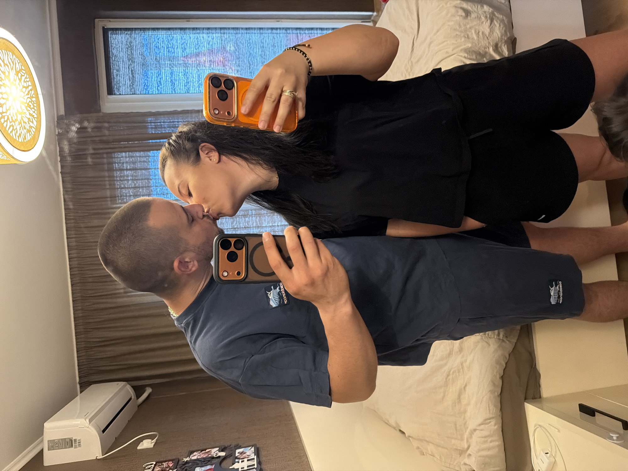 A man and a woman are taking a mirror selfie in what appears to be a bedroom. The man, on the left, is wearing a dark blue t-shirt and shorts. The woman, on the right, is wearing a black t-shirt and shorts. Both are holding smartphones to take the picture, and they are kissing. In the background, there is a bed, curtains, an air conditioning unit, and a light fixture. The location is described as a bedroom in Burgas, Bulgaria.