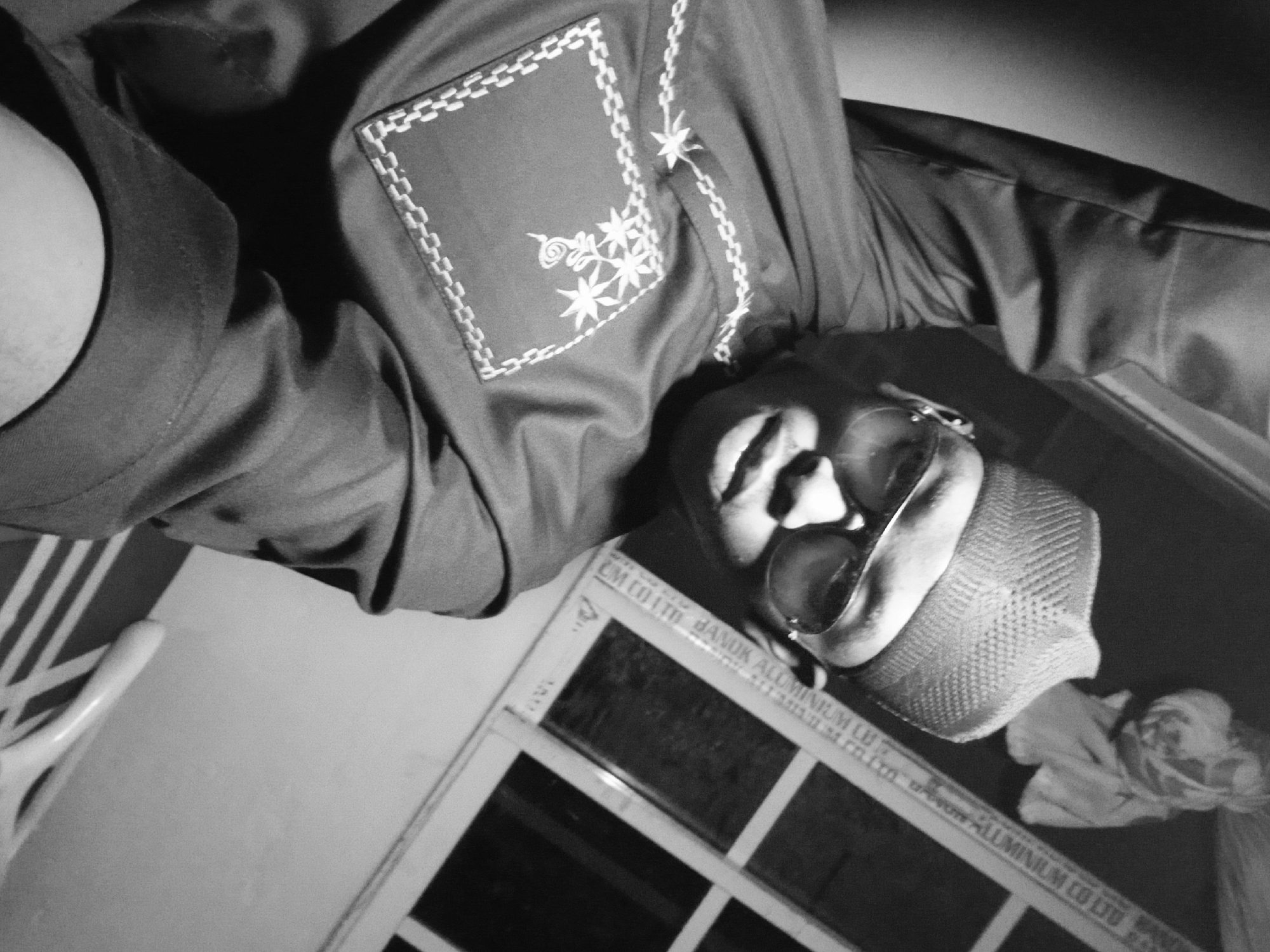 The image is a low-angle, black and white shot depicting a person wearing sunglasses and a patterned kufi cap. The individual is wearing a dark-colored shirt with a decorative, square pocket featuring embroidered floral designs. The scene includes a window frame labeled "CM CO LTD HANOK ALUMINIUM CO LTD," suggesting the location is Lagos, Nigeria, due to the company's presence there. A partially visible white object, possibly fabric, is nearby. The photograph appears to have been taken indoors.
