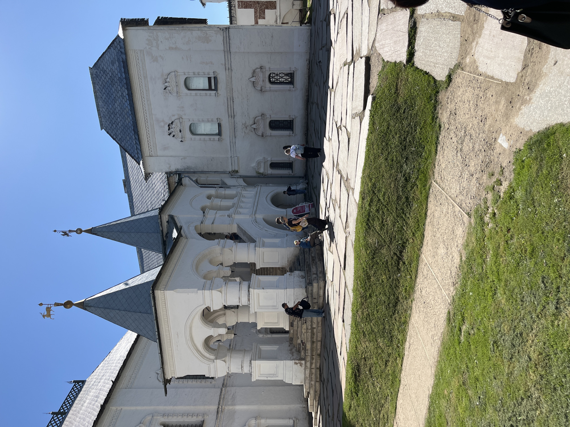 This image depicts the exterior of a white stone building in Novo-Nikolskoye, Russia, under a clear blue sky. The building features traditional Russian architecture with multiple steeply pitched grey roofs topped with decorative weather vanes shaped like stags. A prominent white porch with arched colonnades leads up to the entrance. Several people are visible in the scene. Two individuals stand near the entrance of the porch, one holding a bag. Further to the right, a group of three people are walking on a paved courtyard. Another person is seated on a bench near the porch. The ground is covered with large, irregular flagstones interspersed with patches of green grass. The scene appears to be a sunny day, with clear lighting and some shadows cast by the building and the people.