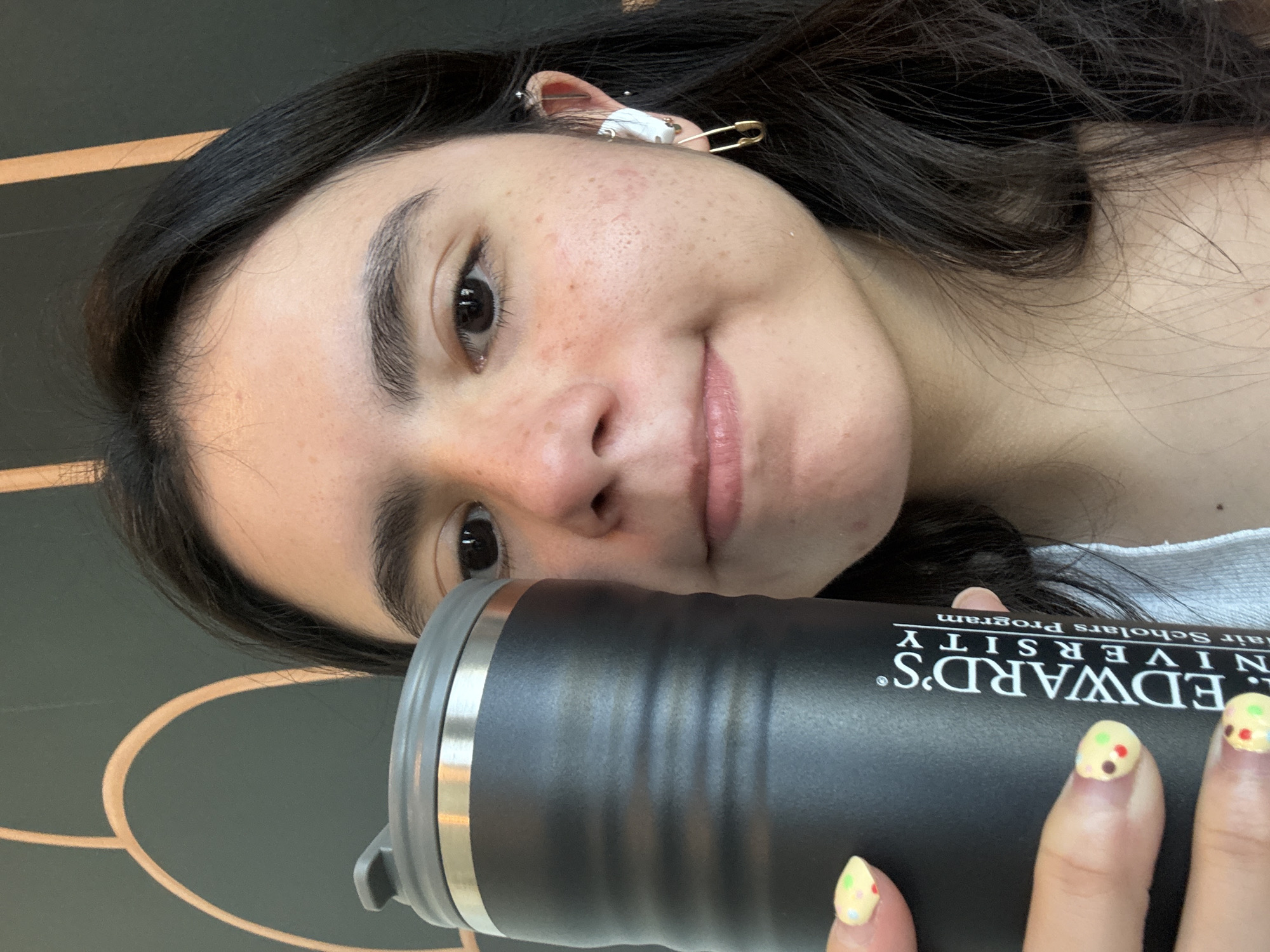 A young woman is holding a black tumbler with the inscription "St. EDWARD'S UNIVERSITY Hainair Scholars Program." She has dark hair, light skin with some freckles, and is wearing an earbud in her left ear, secured with a silver safety pin. Her nails are painted pale yellow with colorful dots. The background is blurred but appears to be a dark surface with a light-colored circular design. Considering the "St. Edward's University" inscription, the location is likely in or around the St. Edward's University campus in Austin, Texas.