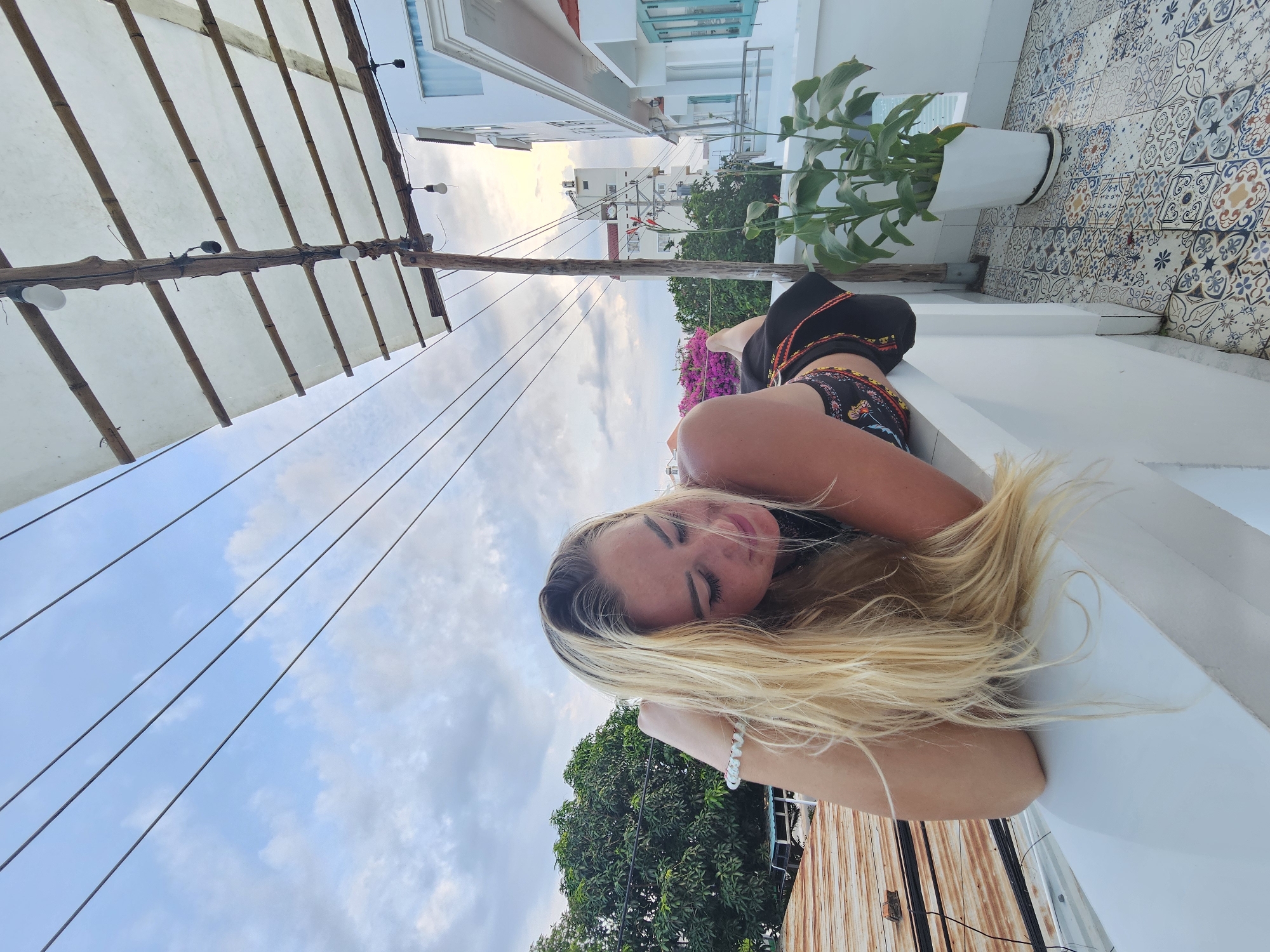 A blonde woman lies on a white railing outdoors. She is wearing a black top with a colorful patterned waistband. Her head is resting on her arm, and her eyes are closed. Behind her, a tree with green leaves is visible, as are several buildings and a clear sky with clouds. The balcony has a tiled floor and a potted plant. The scene appears to be a hotel balcony in Phu Thuy Ward, Vietnam.