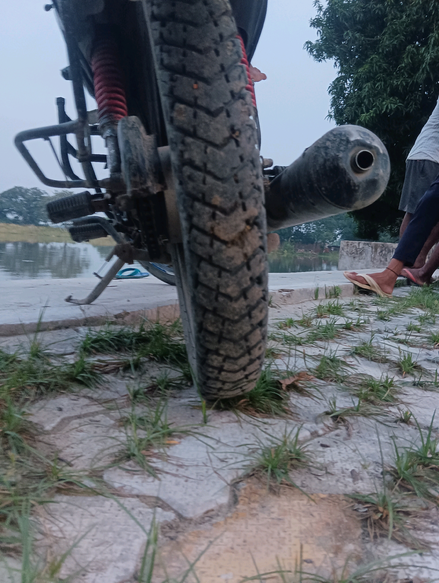 A low-angle, ground-level shot captures the rear tire and exhaust of a motorcycle parked outdoors. The motorcycle's rear shock absorber, with its red spring, is visible. To the right, two individuals are seated, one wearing a white t-shirt and grey shorts, and the other in dark pants. Their legs are visible, and one person wears a light brown flip-flop. The ground is paved with concrete, with grass growing through the cracks. In the background, a body of water is present, with trees and foliage lining the opposite bank. The sky is overcast. The location is identified as being in Tilkar, India.