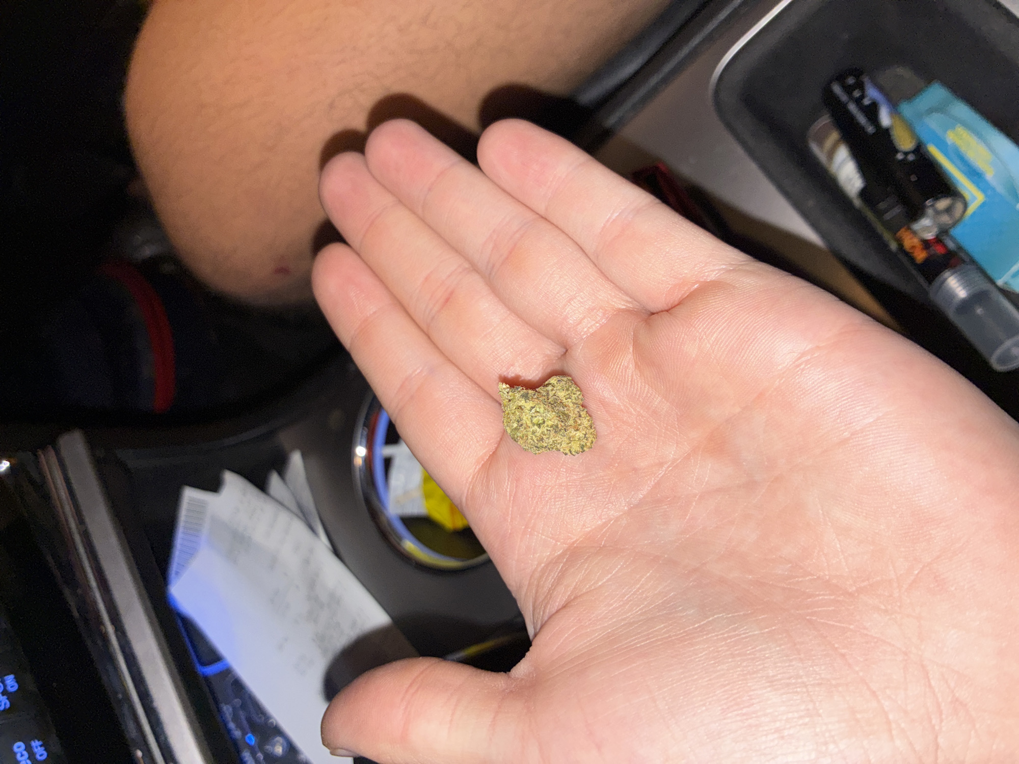 The image captures a person's open palm holding a small, green bud, likely cannabis, against the backdrop of a car interior at night or in a dimly lit setting. The hand, with pale skin and visible palm lines, is the central focus. In the blurred background, another person's arm or shoulder with a small red blemish is partially visible.
The scene takes place inside a vehicle, indicated by various elements. To the right of the hand, a black tray or compartment holds a black vape pen with colorful accents and a blue package. Below the hand, crumpled paper receipts are tucked into a slot, some displaying barcode patterns. Car controls are also visible, featuring blue backlighting and text such as "NO ECO OFF" and "ECO." The lighting suggests an indoor or nighttime environment. No emotions are discernible. The location is specified as Huixquilucan de Degollado, Mexico.