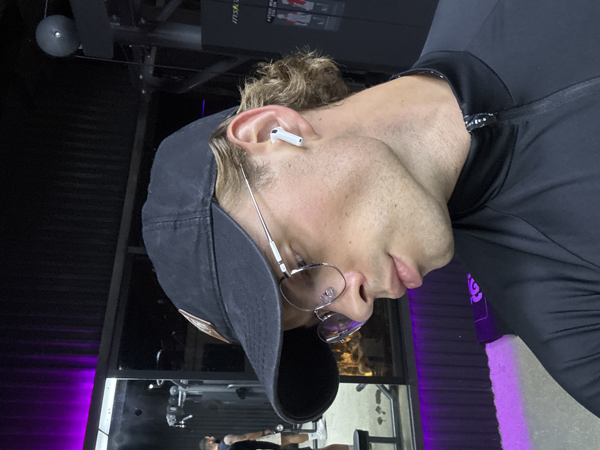 A man is shown from the head down to just past the shoulders, wearing a black baseball cap, glasses, a white Airpod in his left ear, and a black athletic shirt. He is in a gym with purple LED lighting. A weight machine can be seen in the background. There is no indication of the location in the city of Canggu, Indonesia.