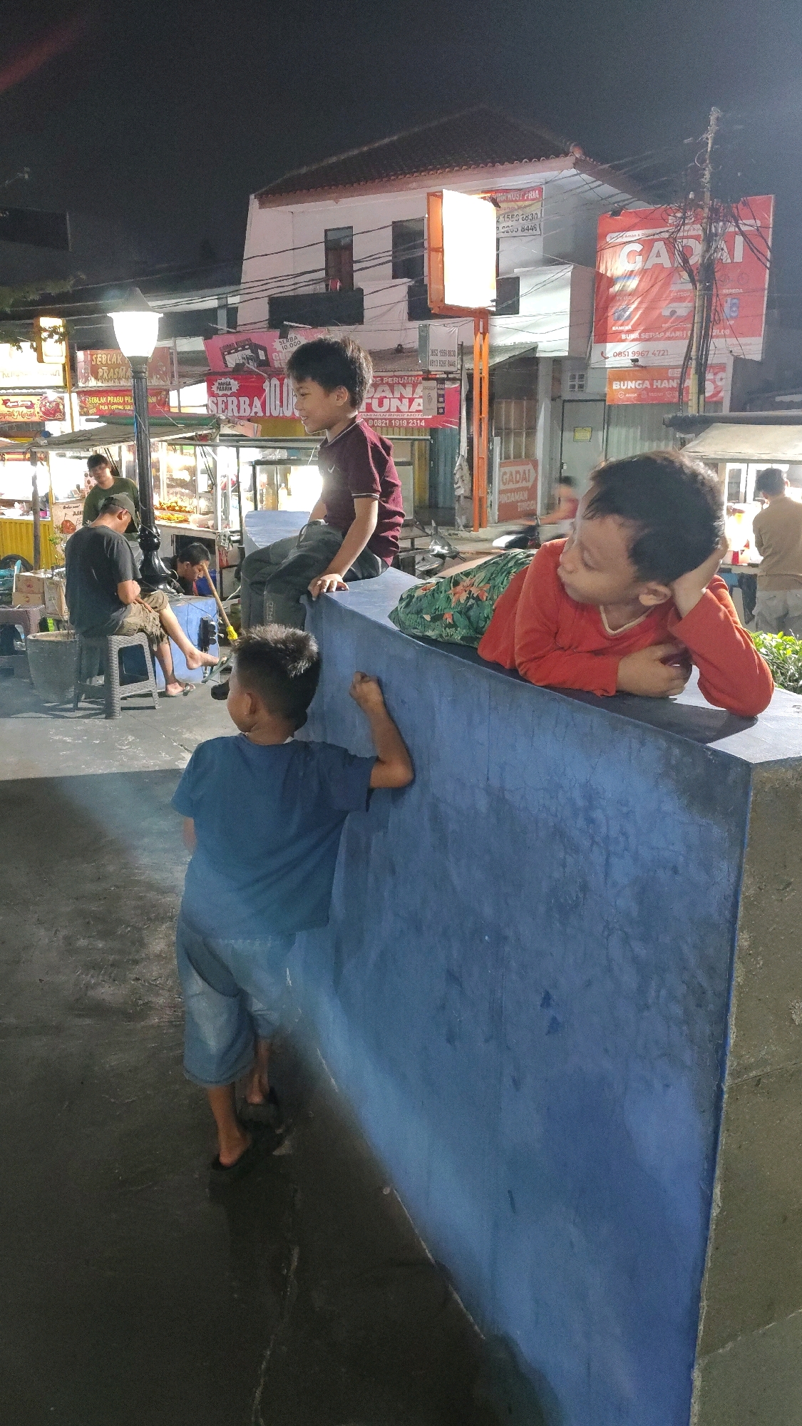 The image depicts an outdoor scene at night, likely in a public area with food stalls. The background features a building with multiple stories and signs, including one that reads "GADAI". There are also illuminated signs with text and what appear to be food vendors. In the foreground, two young boys are the central subjects. One boy, wearing a blue t-shirt and shorts, is standing with his back to the camera, leaning against a blue concrete wall and looking towards the right. The other boy, wearing an orange long-sleeved shirt, is lying on the same blue concrete structure, resting his head on his hand and looking to his left. Further in the background, other individuals are visible, some seated and others standing, near the food stalls. The ground appears wet, reflecting the ambient light. The presence of signs with Indonesian text, such as "GADAI" (pawnshop) and "SEBLAK PRASMAN" (a type of Indonesian food), strongly suggests the location is in Indonesia, and considering the visual cues, potentially in Tangerang, given the style of shops and signage commonly found in that area.