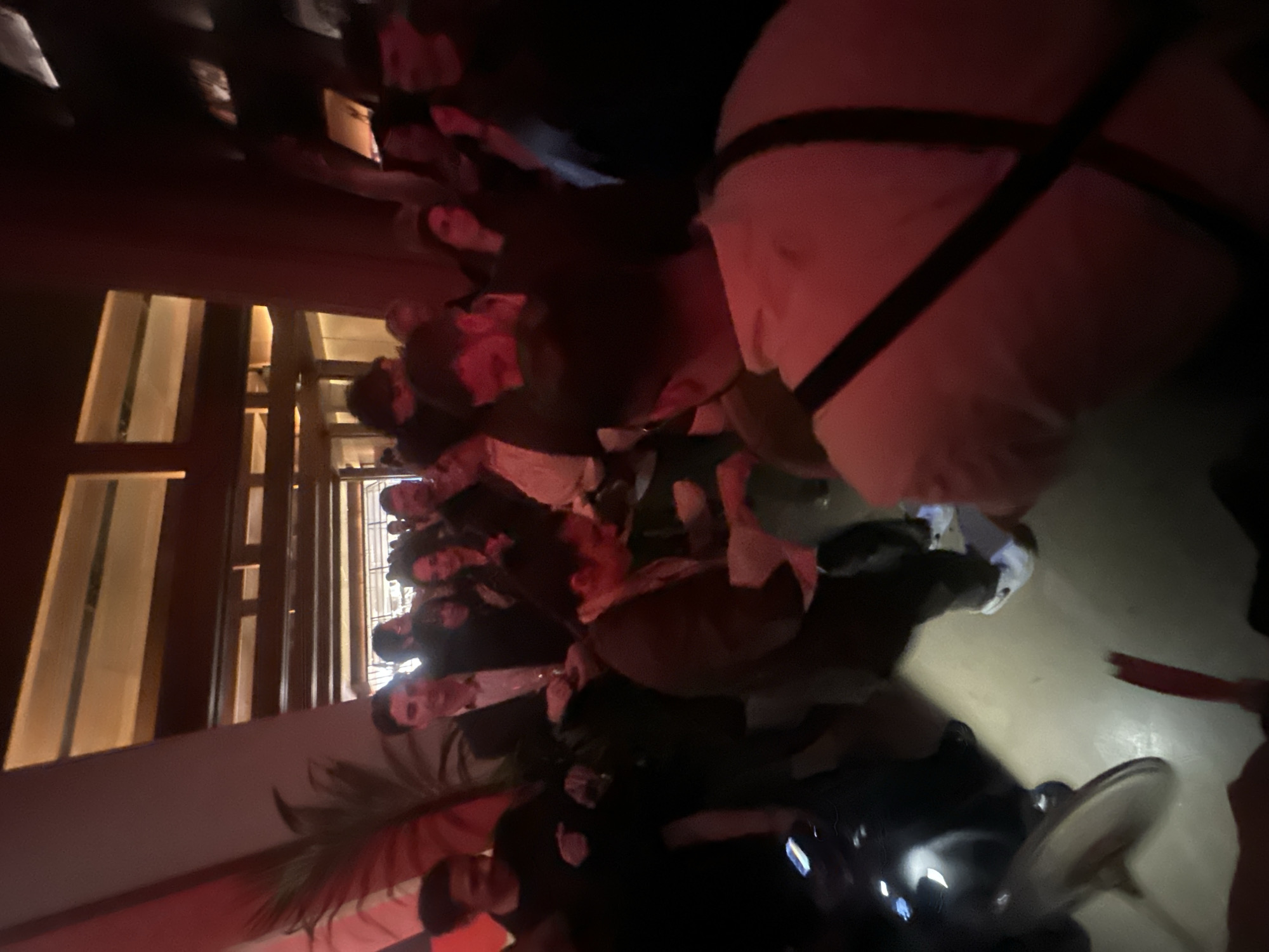 This image captures a crowded indoor social gathering, likely a club, bar, or event space, with numerous individuals, predominantly young adults, populating the scene. The perspective is angled, making the floor appear as a wall and giving a dynamic, almost disorienting, view of the activity.
**Subjects:** The primary subjects are the many people standing close together, indicating a lively, packed venue. Their faces are often partially obscured, out of focus, or dimly lit, making individual identification difficult. They are dressed in casual to semi-formal attire. Objects visible include drinks in hands, a glimpse of white athletic footwear, and a smartphone screen emitting light in the lower right corner. Architectural features like dark wooden or metallic frames and vertical, illuminated panels (possibly windows or decorative partitions) are seen on the left side of the frame. A green, leafy plant adds a decorative element in the bottom left.
**Activity or event taking place:** People are gathered in a social setting, standing, mingling, and presumably conversing. The atmosphere appears to be one of an active party or event.
**Notable details:** The scene is characterized by dim, atmospheric lighting. Red-toned artificial lights cast a warm, sometimes intense, glow on people and surfaces, creating a vibrant mood typical of evening social venues. Brighter, more neutral light emanates from the background architectural features on the left. The time of day is clearly night or late evening due to the artificial illumination. There are no explicit location cues within the image to specify a particular spot in London, nor is any text visible. Emotions are difficult to discern with certainty due to the image quality and distance, but the overall impression is one of engagement and enjoyment among the crowd.