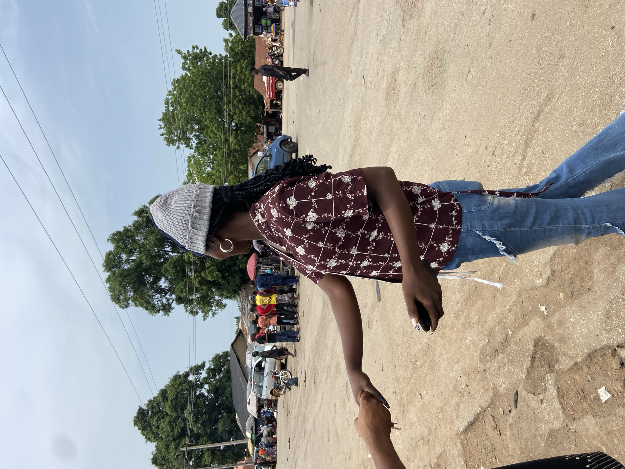 The image depicts a woman in Mararaba, Nigeria, standing on a sandy, outdoor street surface. She is wearing a gray beanie, a patterned maroon shirt, and blue jeans with frayed hems. She is holding a black mobile phone in her right hand and reaching out to shake hands with another person whose arm is visible in the foreground. In the background, there is a small crowd of people, vehicles, buildings, trees, and overhead power lines. A person can be seen walking down the street.