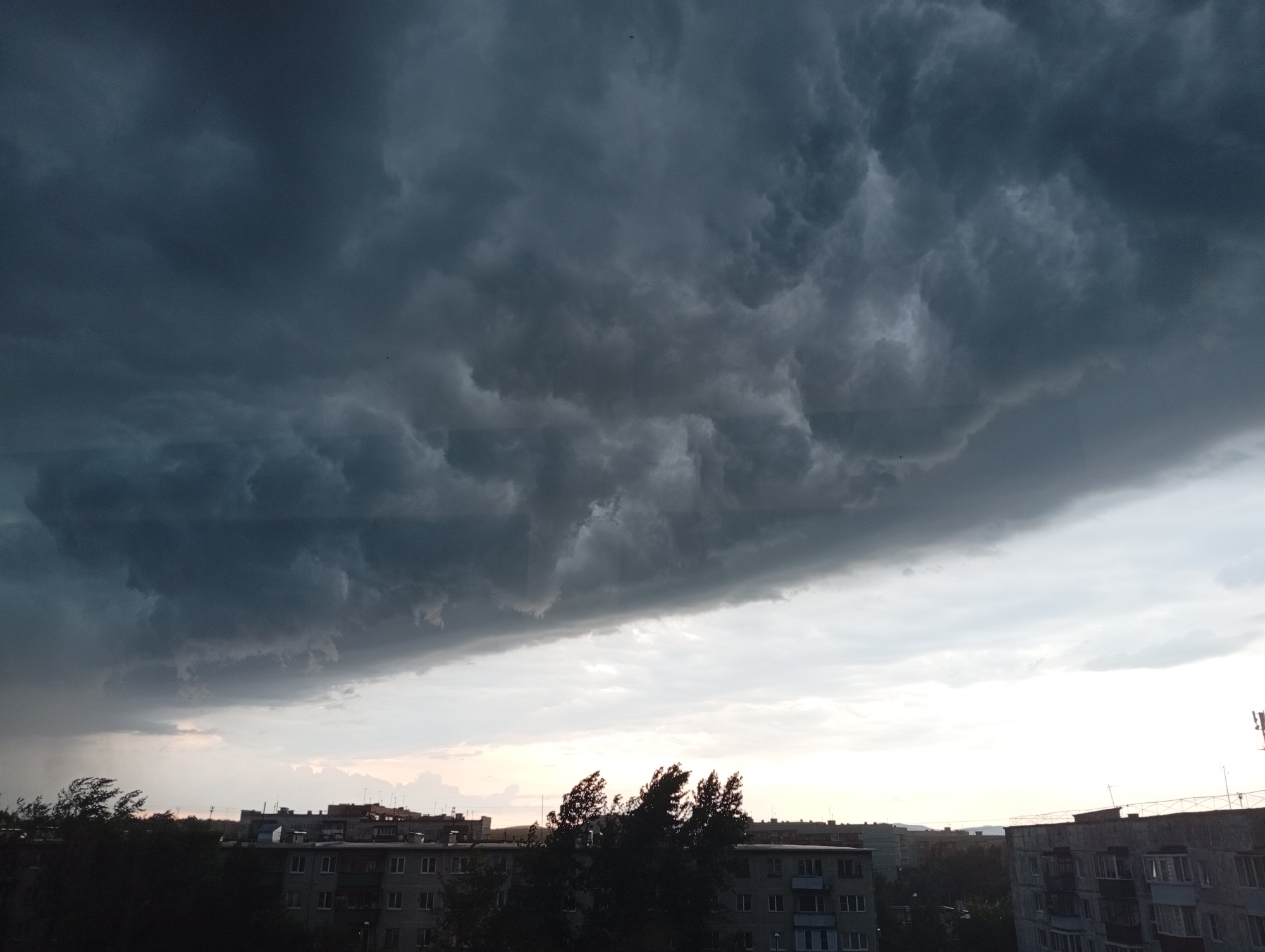 This image captures a dramatic sky over the city of Kyshtym, Russia, likely taken from an elevated vantage point. The sky is dominated by dark, ominous storm clouds that fill the upper two-thirds of the frame. These clouds have a turbulent, textured appearance, with deep shadows and lighter, swirling patterns. Below the storm clouds, a sliver of lighter sky is visible, transitioning to a hazy, pale sunset glow near the horizon.
In the foreground, the silhouettes of apartment buildings are visible, characteristic of urban Russian architecture. These buildings appear to be several stories high, with numerous windows and balconies. Dark trees, their branches appearing windswept, are scattered in front of and between the buildings, suggesting strong gusts of wind accompanying the approaching storm. The overall impression is one of an impending weather event, with the city bracing for a storm. There are no visible people or animals in the frame. The scene is devoid of any discernable actions or interactions.