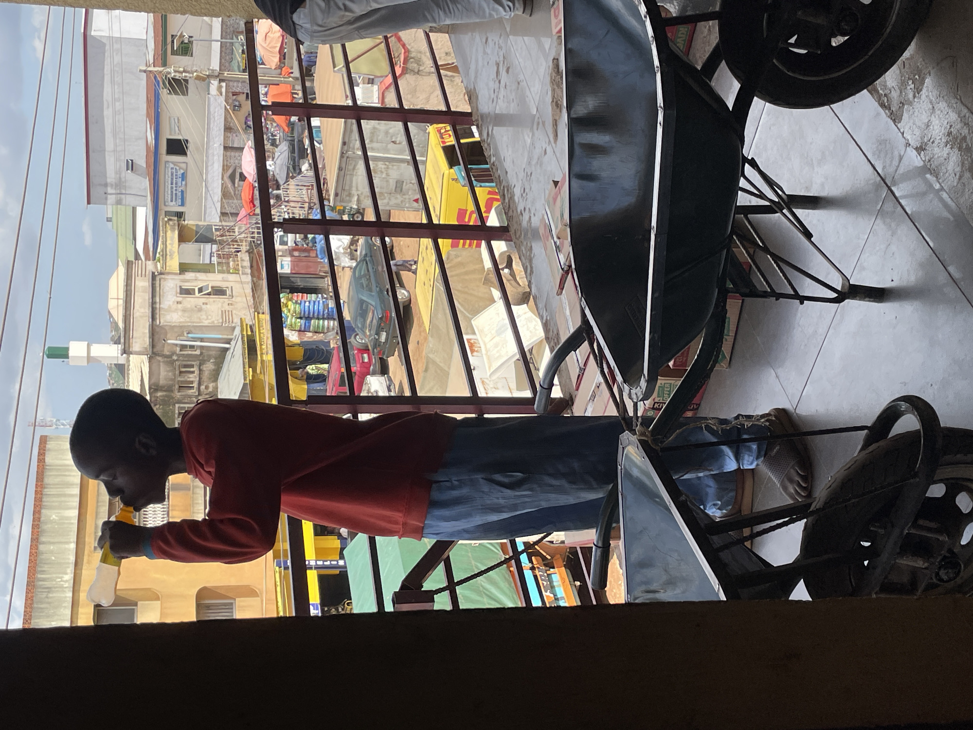 The image shows a young boy drinking from a bottle while standing on a tiled floor, possibly a balcony or walkway. He is wearing a red long-sleeved shirt and blue jeans. Behind him, a wheelbarrow with a large dark basin and a wheel is visible. The wheelbarrow appears to be metallic and possibly used for construction or transport.
In the background, there are buildings with various architectural styles, suggesting an urban or semi-urban setting. Some buildings have balconies and windows, and there are power lines running across the scene. There are also glimpses of what appears to be a market or a busy street, with stalls and people. A yellow truck with "DHL" branding is partially visible. The sky is overcast, indicating a cloudy day.
The boy's posture suggests he is focused on drinking from the bottle. There are no strong emotional cues visible. The overall scene appears to be a snapshot of everyday life in a Nigerian location, possibly Jos, given the context. The time of day is difficult to determine precisely due to the overcast conditions, but it appears to be daytime.