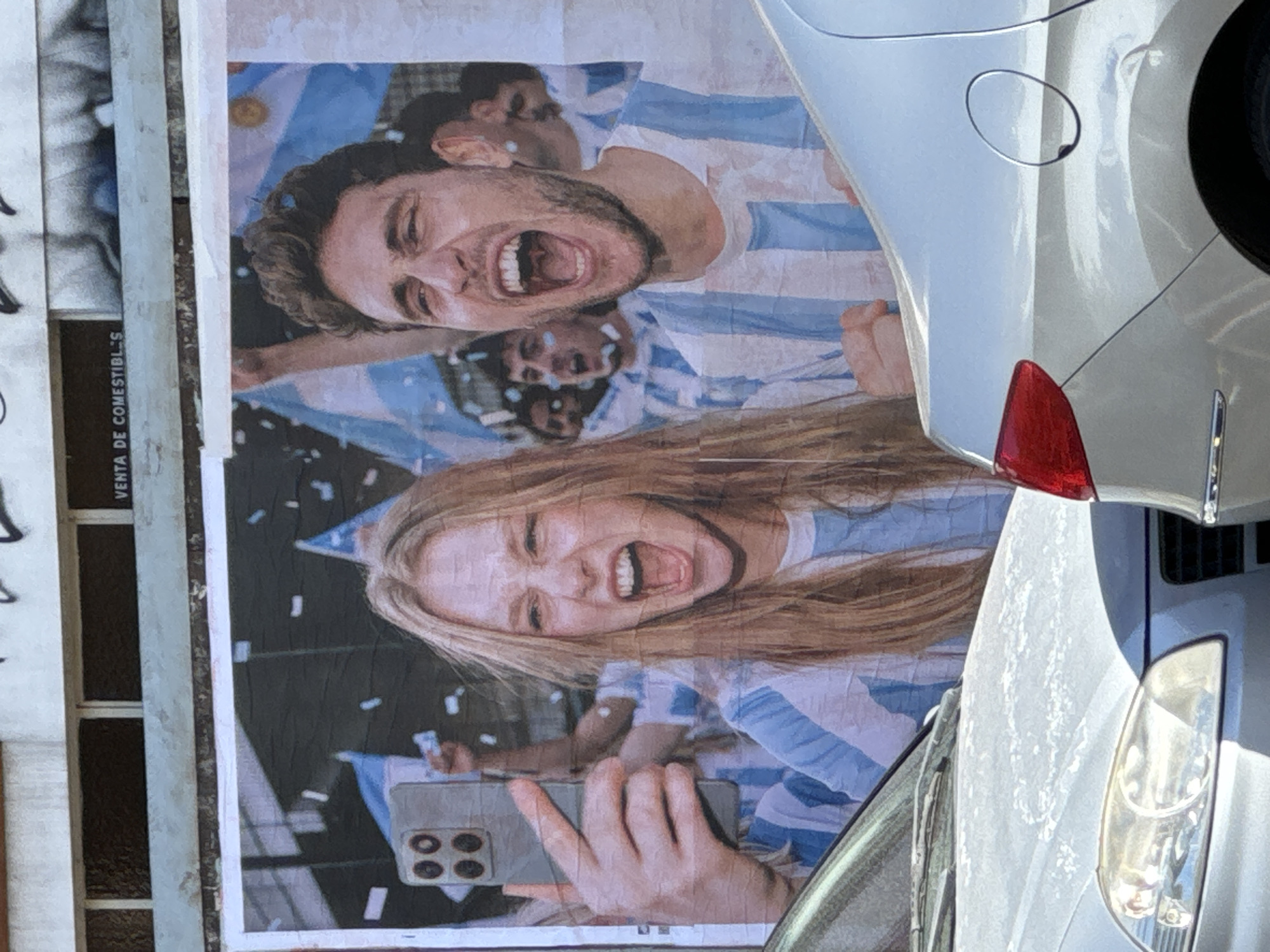 The image shows a poster in Buenos Aires, Argentina, featuring two people cheering, likely celebrating a victory. They are wearing Argentina's national soccer team's striped blue and white jerseys. The woman is holding a smartphone and taking a selfie. The poster is attached to a wall next to a storefront and partially obscured by a parked silver car. A sign with the text "VENTA DE COMESTIBLES" (sale of edibles) is visible on the storefront.