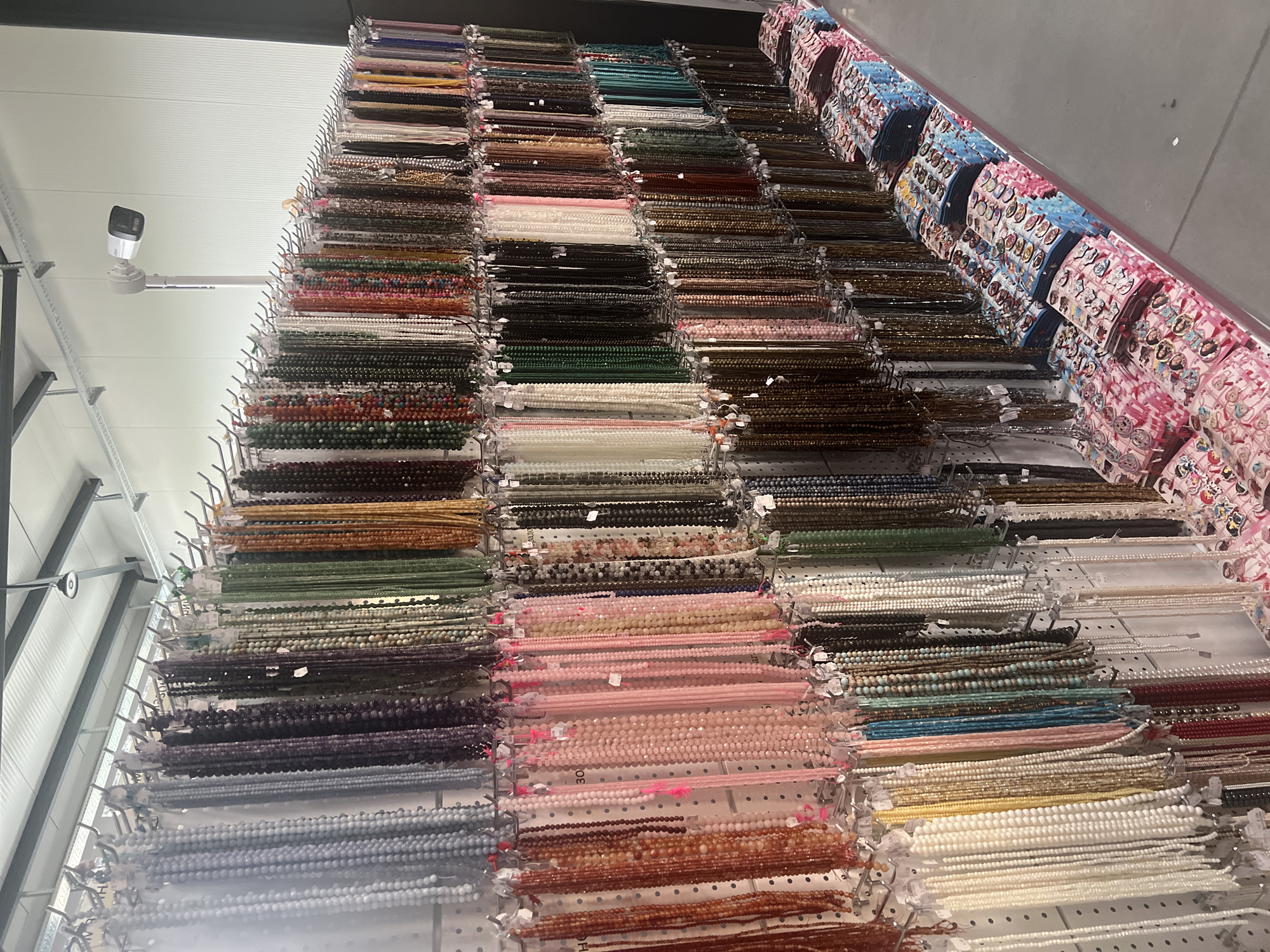 The image shows a wall display of numerous strands of beads of various colors and materials in what appears to be a retail or craft store. The beads are arranged in rows, hung on metal racks with clear plastic dividers. To the right of the frame are packaged items with cartoon-like designs on them, possibly indicating children's merchandise.
While the location is unknown without more specific information, the prompt mentions that the location may be in the city of Burgas, Bulgaria. The presence of surveillance camera may suggest that the location is a commercial retail location.