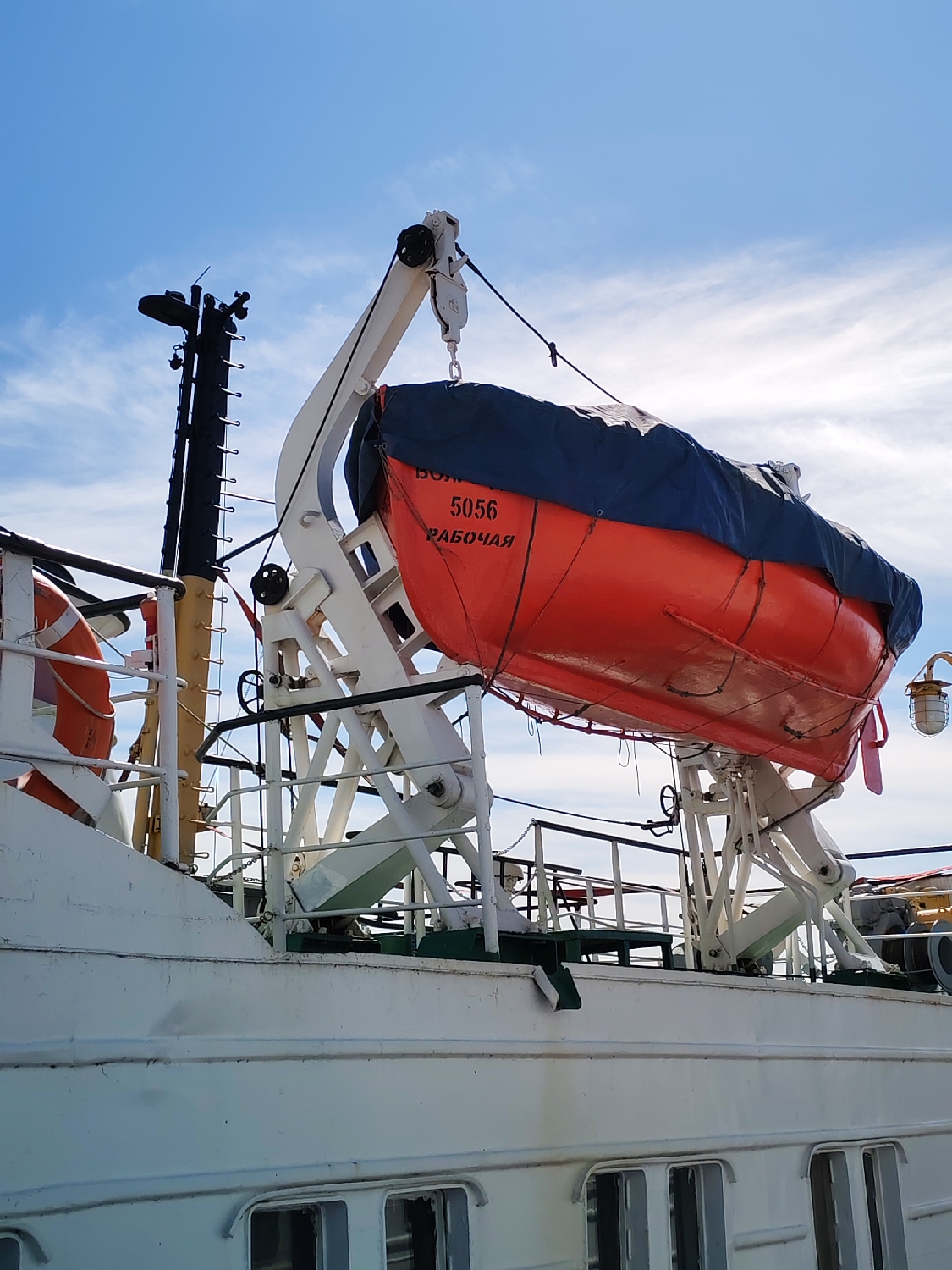 The image shows a detailed view of the upper deck and side of a white ship, likely a river vessel, against a bright blue sky dotted with scattered white clouds. The central feature is a vibrant orange lifeboat, partially covered with a dark blue tarpaulin, securely stowed in its white davits.
The lifeboat is suspended from a robust white crane-like structure equipped with a pulley system and ropes. On its side, black lettering clearly spells out "ВОЛГА" (Volga), followed by the numerical identifier "5056", and below that, the word "РАБОЧАЯ" (Rabochaya), which translates to "working" or "workboat". This indicates the vessel is a workboat, possibly named "Volga 5056".
To the left of the lifeboat, a tall, black mast with various antenna-like structures and ladders rises towards the sky. Part of an orange and white lifebuoy is visible attached to the ship's railing in the lower-left section. The ship's hull and superstructure are painted white, showing signs of wear and multiple rectangular windows along the lower deck. Visible deck railings and hand wheels for operating equipment are also present.
The scene takes place during bright daylight, suggested by the clear illumination and the blue sky, indicating fair weather conditions. There are no people visible, and no active event is occurring, with the lifeboat appearing to be in its standard stowed position. The provided context places the location in Rybinsk, Russia, a city on the Volga River, which aligns perfectly with the "Volga" inscription on the lifeboat. The overall impression is of a well-equipped and maintained working vessel.