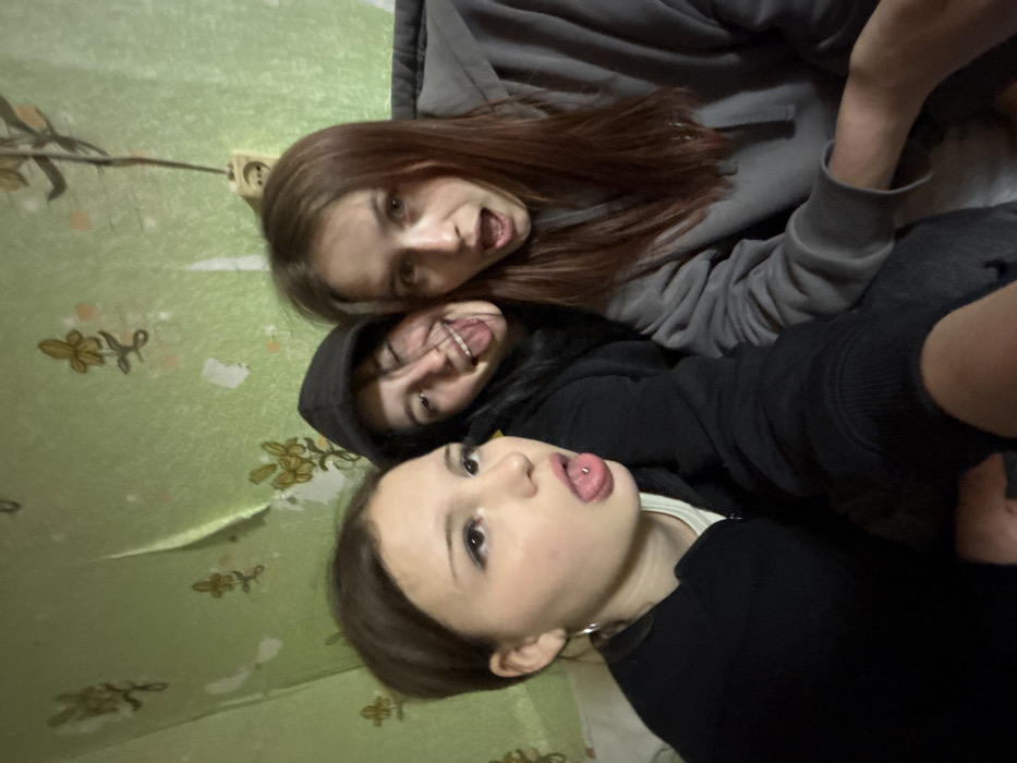 An indoor, close-up photograph depicts three young females posing playfully within a residential setting in the city of Фоки, Russia. The background features a light green wallpaper with a repeating pattern of stylized golden-brown flowers, exhibiting visible wear, tears, and peeling sections. An exposed electrical outlet with an attached dark wire is present on the upper right portion of the wall.
From left to right:
1. A female with dark hair and visible eyeliner wears a silver hoop earring on her left ear. She is seen sticking out her tongue, which displays a barbell piercing. She is dressed in a black zipped jacket over a light-colored top.
2. A female with dark hair wears a dark hooded sweatshirt with the hood pulled up. She is winking her right eye and sticking her tongue out.
3. A female with long, reddish-brown hair wears a grey hooded sweatshirt. Her mouth is open in a wide, expressive manner.
All subjects are looking towards the camera, indicating a posed shot, likely a selfie, characterized by informal and jovial expressions.