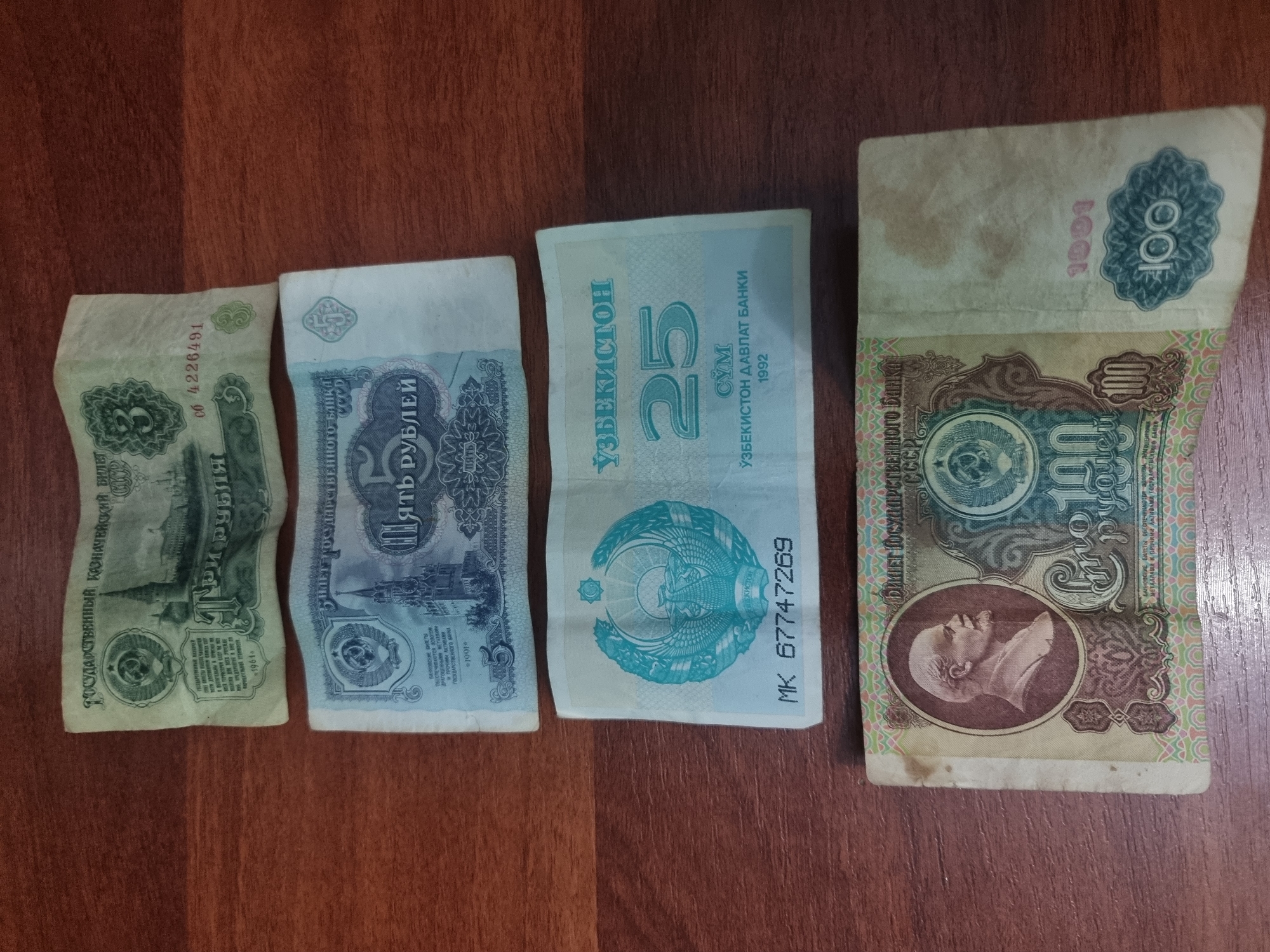 The image shows four pieces of currency laid out on a wooden surface. The currency appears to be from different countries and eras.
From left to right, the first bill is green and has text that appears to be in Cyrillic script. Visible text includes "ГОСУДАРСТВЕННЫЙ КАЗНАЧЕЙСКИЙ БИЛЕТ" and "1961". It also has a serial number "CÓ 4226491".
The second bill is light blue and features a building and Cyrillic text. Prominent text includes "ПЯТЬ РУБЛЕЙ" and "ГОСУДАРСТВЕННОГО БАЙНА".
The third bill is light blue and has text in both Cyrillic and Latin scripts. It prominently displays "25" and "CYM" in large font, along with "ЎЗБЕКИСТОН" and "ЎЗБЕКИСТОН ДАВЛАТ БАНКИ 1992". A serial number "MK 67747269" is also visible. This bill is from Uzbekistan.
The fourth and largest bill is light brown and features a portrait of a man, likely a political figure. It displays "100" in large font and has Cyrillic text including "ГОСУДАРСТВЕННОГО БАНКА СССР" and "1991". It also has text that includes "БАНКОВСКИЕ БИЛЕТЫ ОБЕСПЕЧИНАЮТСЯ ЗОЛОТОМ".
The wooden surface suggests an indoor setting, likely a table. The lighting is even, indicating no specific time of day or weather conditions are evident. The objects are the sole focus of the image.