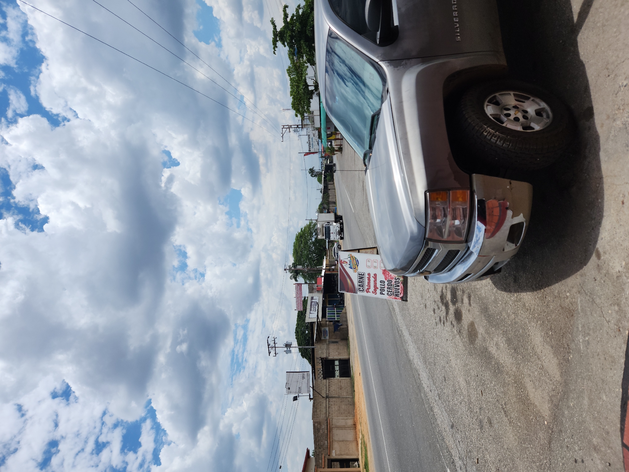 The image depicts a street scene in Tinaquillo, Venezuela. A silver Chevrolet Silverado truck is parked on the right side of the road. A sign advertising meat products (carne, pollo, cerdo, huevos) is visible in front of a building, likely a butcher shop or grocery store. Power lines run parallel to the road against a backdrop of a cloudy sky. Other buildings and trees are visible along the street. Other cars can be seen in the distance. The road surface is paved.