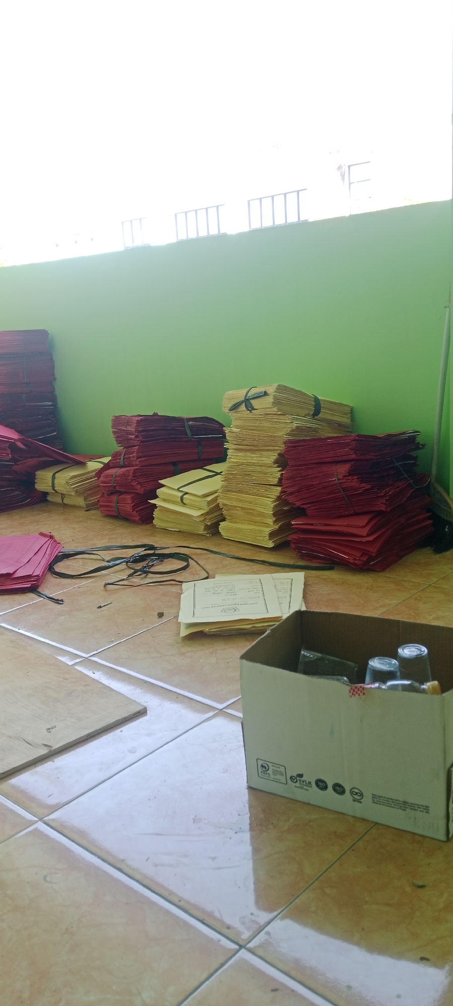 This image depicts a storage area, likely in Tuban, Indonesia, filled with stacks of red and yellow paper, possibly for crafting or manufacturing purposes. The papers are neatly bundled with dark ties. In the foreground, a cardboard box contains several glass bottles and cups. The floor is tiled with a light brown pattern. A green wall serves as the backdrop, with a window or opening at the top showing a bright, possibly outdoor, scene with some railing-like structures. The overall impression is one of an organized workspace or storage facility.