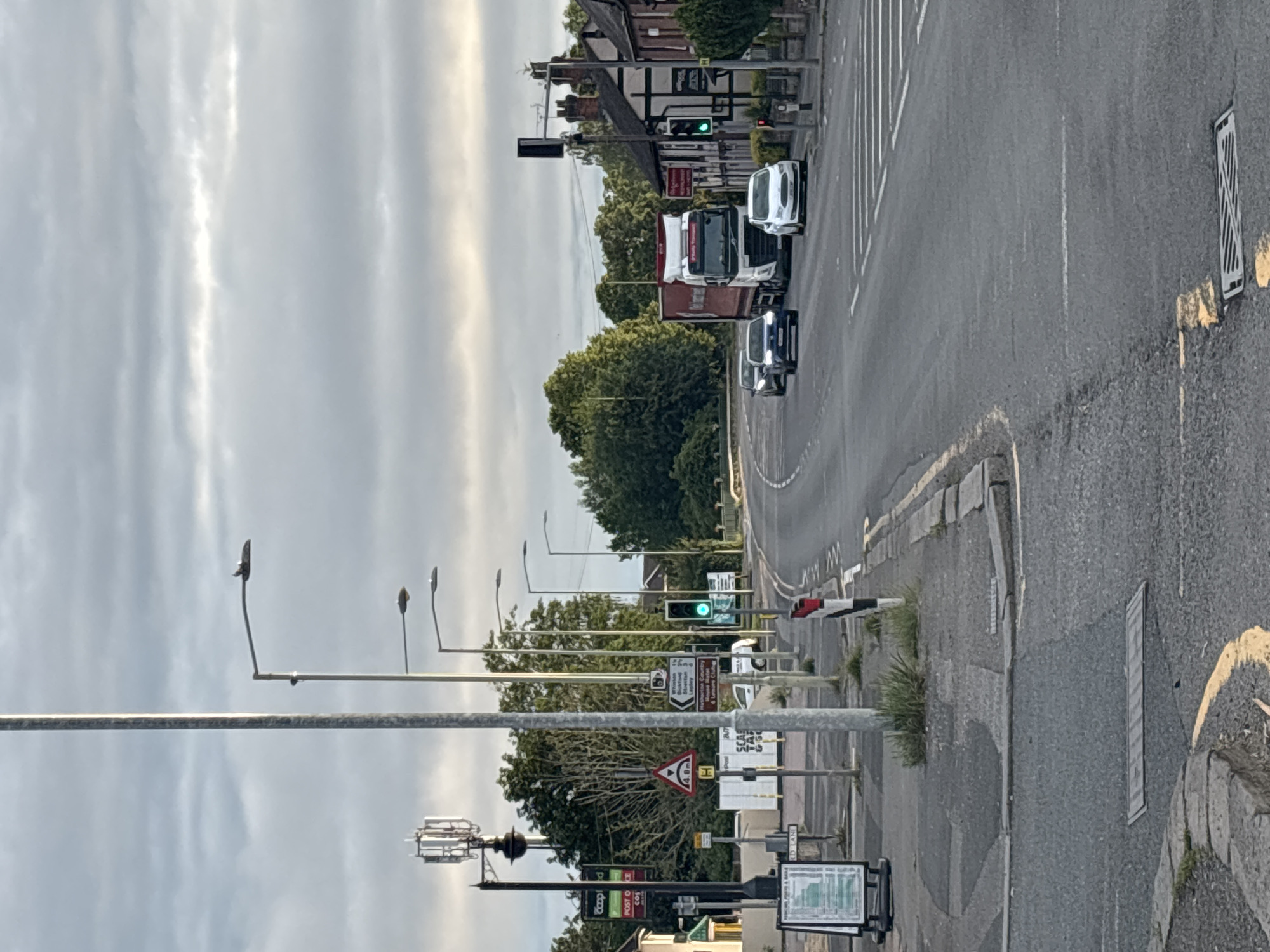 The image depicts a street scene in South Staffordshire, United Kingdom. A grey, overcast sky with textured clouds forms the backdrop. In the foreground, a two-lane road runs horizontally, with white road markings and a yellow curb. A number of streetlights are visible along the side of the road, as well as a tall pole with an antenna at the top.
Several vehicles are present: a white articulated lorry, a white Ford car, a dark blue car, and a second dark blue car further down the road. There is a traffic light that is currently green. On the right side of the road, there are buildings with Tudor-style architectural elements, and dense green trees line the background. Signs for "Whiston, Brocton, Ingestre, Stafford" and a red and white striped barrier are visible. A white pedestrian crossing is marked on the road. On the left side of the road, there is a post box, a "Co-op" sign, and a sandwich board advertising parking facilities. The pavement has some patchy grass growing through it.
