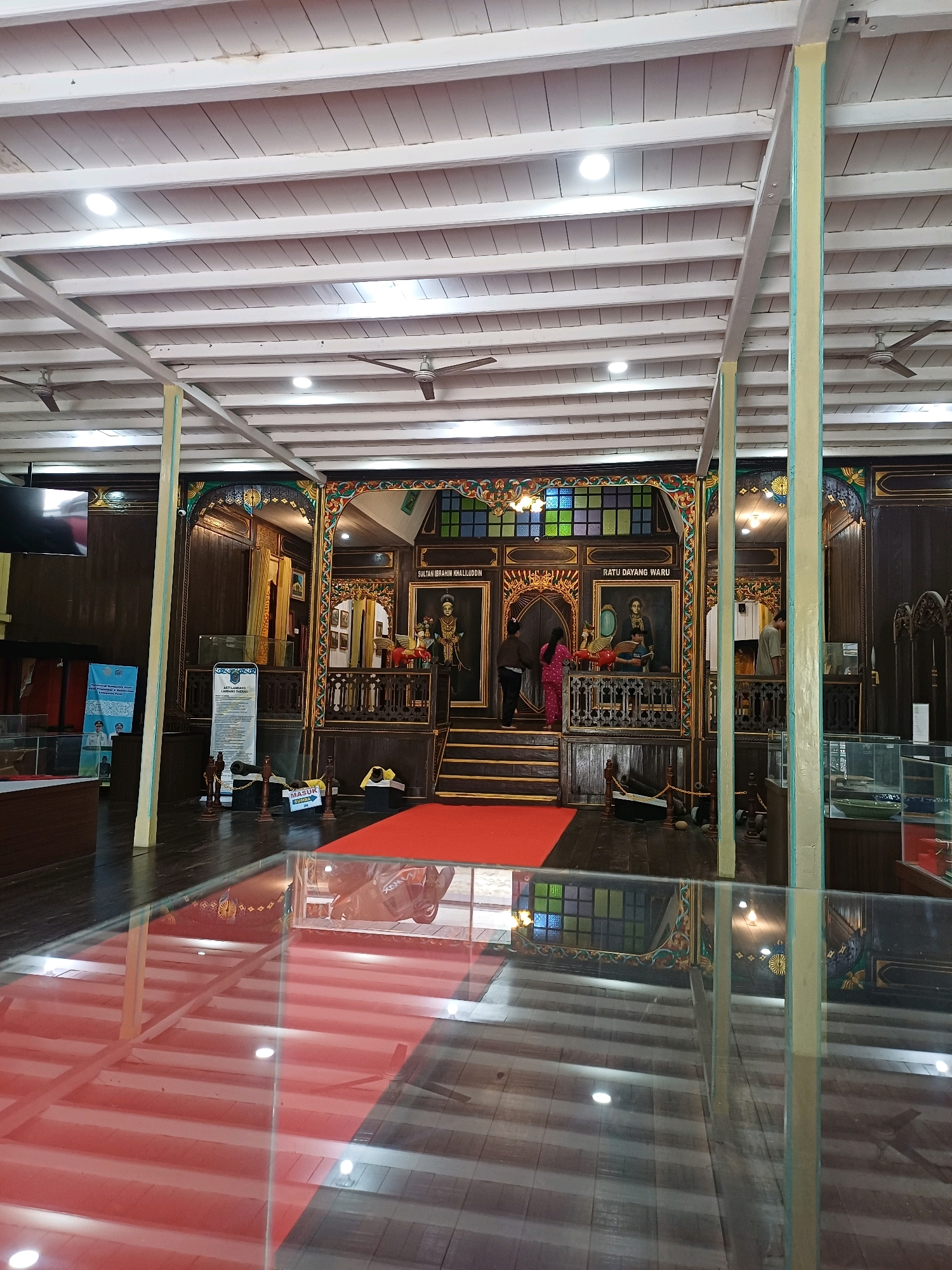 The image shows the interior of a museum in Pasir Belengkong, Indonesia. The museum features traditional Indonesian architecture and artifacts. In the foreground, a glass floor allows visitors to see exhibits beneath. The room is decorated with intricate carvings and colorful stained glass. Several mannequins are displayed, dressed in traditional attire, depicting historical figures. A red carpet leads to a raised platform where more exhibits are presented. Two individuals, a man and a woman, are visible, interacting with the exhibits. The ceiling is made of white wooden beams, and ceiling fans are installed for ventilation. The overall atmosphere is one of cultural preservation and historical display.