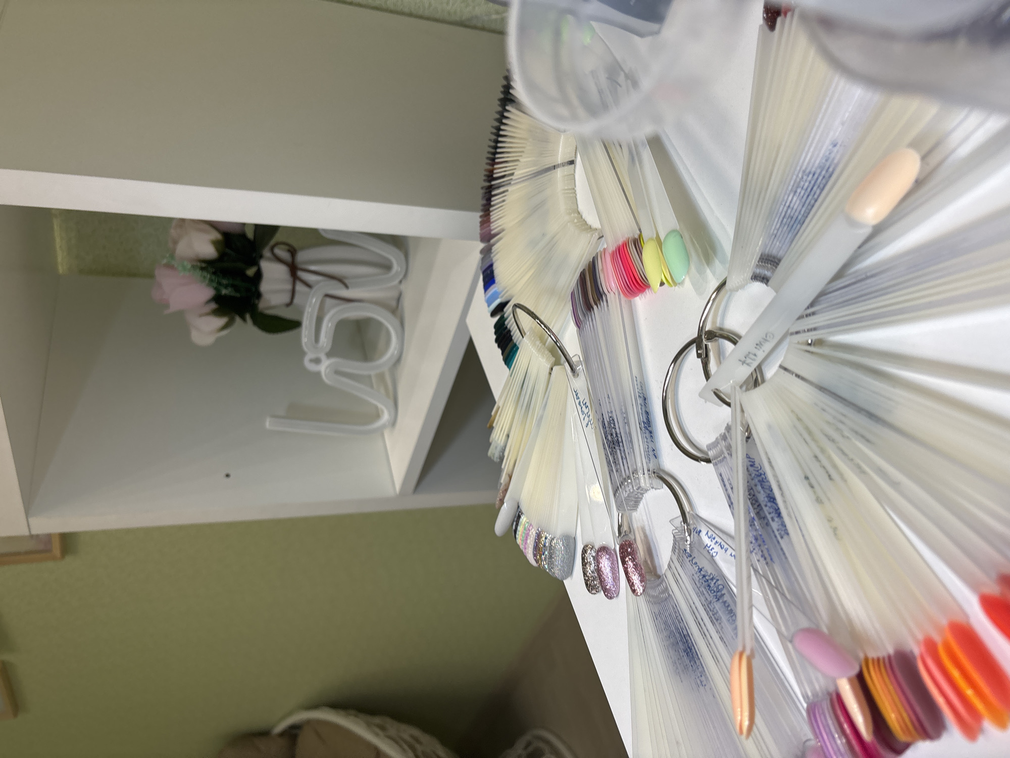 The image shows a brightly lit interior space. In the foreground is a collection of nail polish samples displayed on clear plastic sticks attached to metal rings. The samples are arranged in a fan-like fashion, showing a variety of colors. In the background, there is a white shelf containing a bouquet of pink and white flowers in a vase, along with a decorative white "LOVE" sign. The wall is a pale green color. A part of a wicker basket or chair is visible in the lower part of the image. Based on the context of the prompt mentioning Ферма, Russia, it can be inferred that the image was taken in this location.