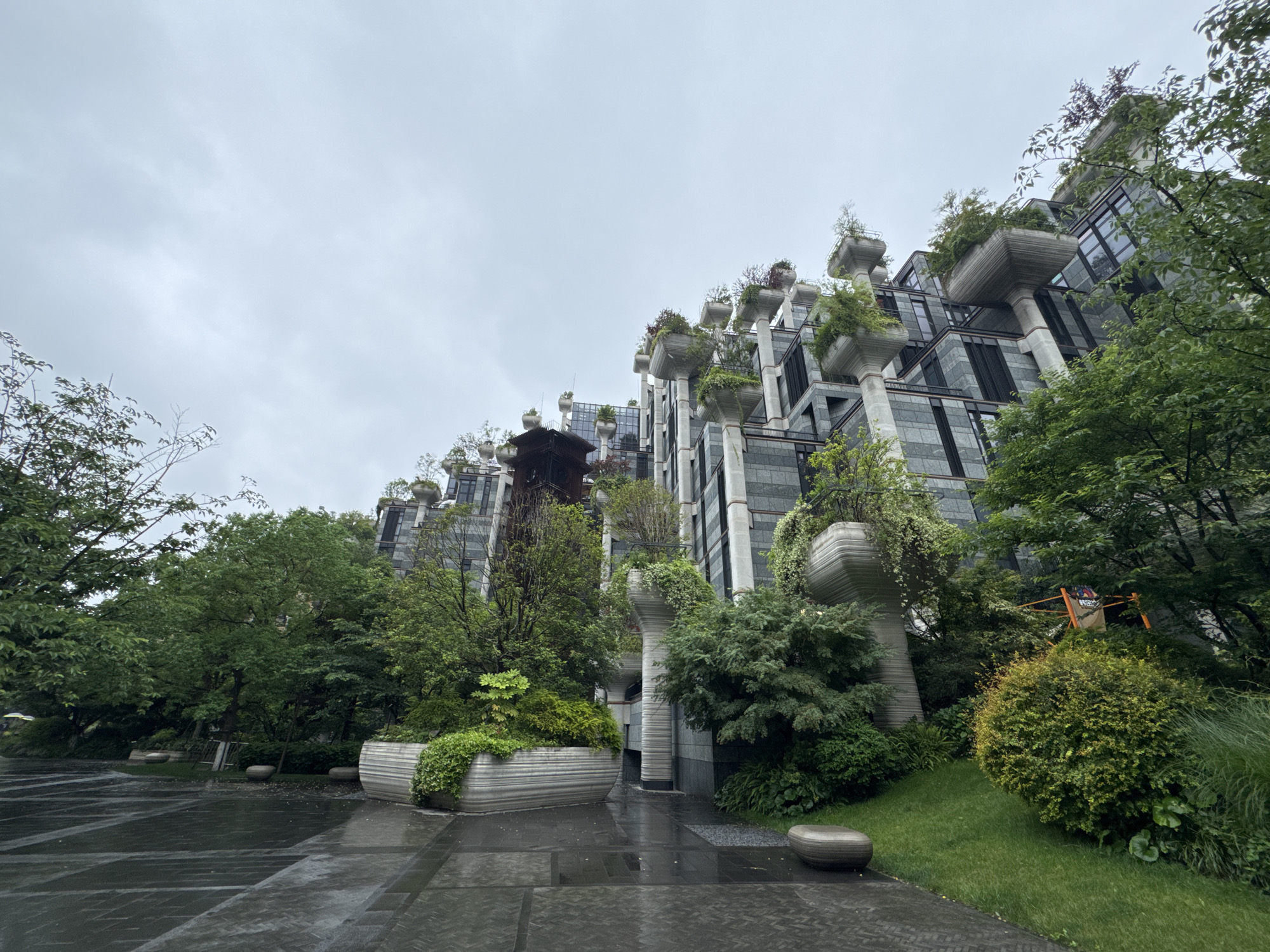 The image shows a low-angle view of the 1000 Trees complex, also known as Moganshan Road, located in Putuo District, Shanghai, China. The complex features modern architecture with a distinctive design. The buildings are characterized by a series of concrete pillars topped with planters containing greenery, creating the impression of trees growing from the structure. The overall design incorporates an abundance of vegetation, with trees and plants surrounding the base of the buildings and on the elevated planters. The weather appears overcast with a gray sky. The ground is wet, possibly from rain. The foreground features a paved area constructed of large rectangular stones, with several raised planters and shrubs nearby. In the midground, a traditional-style Chinese pavilion or structure is visible between the modern buildings. The architectural style is modern with Asian influences. In the distance, scaffolding or construction equipment is visible. There are no people visible in the image.