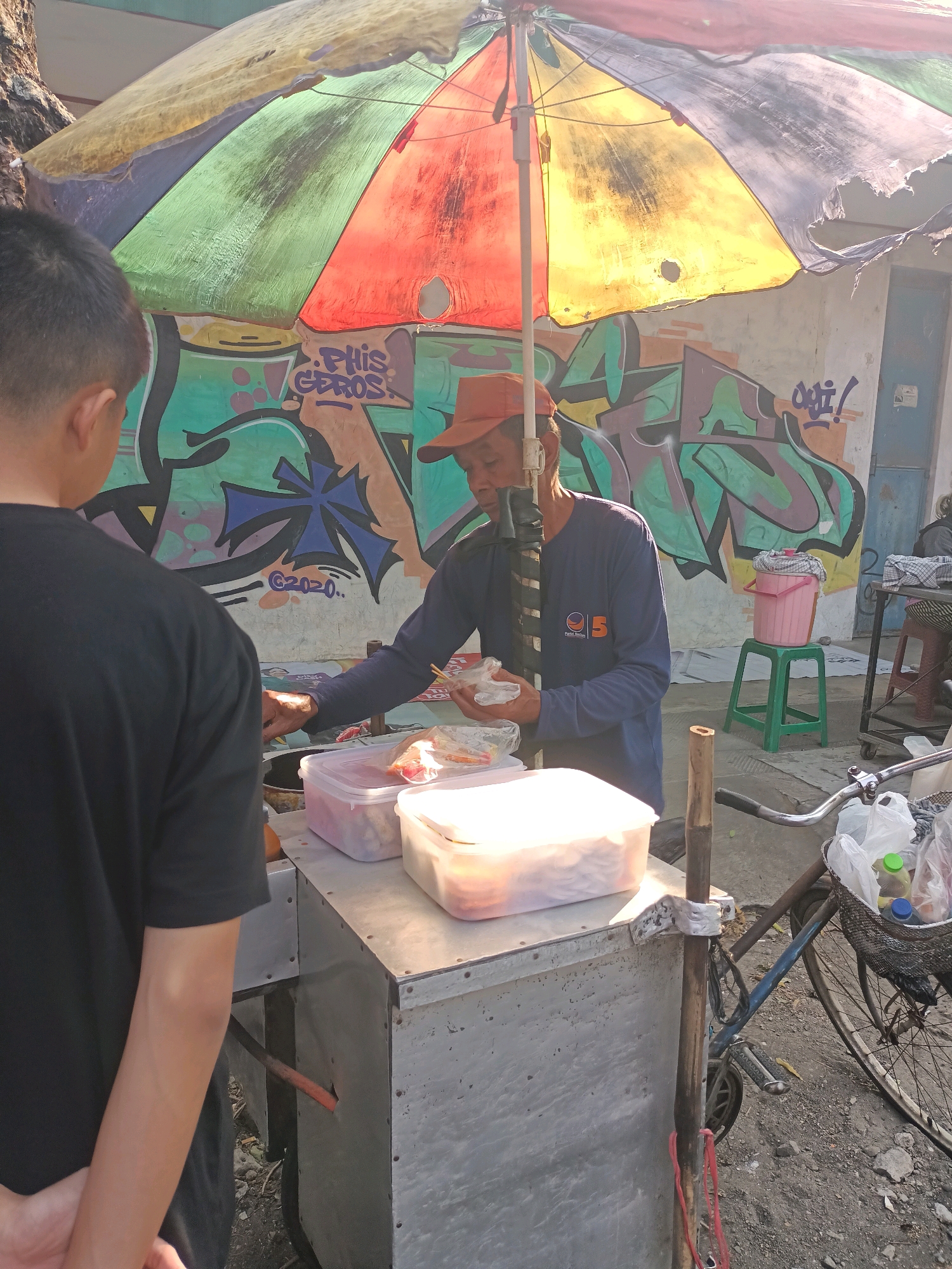 In the city of Pare, Indonesia, a street vendor wearing a red cap and a blue long-sleeved shirt is serving a customer. The vendor is standing behind a food cart, which has several clear containers of food on top. The vendor is holding a packaged food item in their left hand and appears to be handing it to the customer, who is seen from the back wearing a black t-shirt. The food cart is covered by a colorful, tattered umbrella, with panels of red, yellow, green, and blue. Behind them, a wall is covered in graffiti. To the right of the cart, a bicycle is parked, with a basket containing plastic bags and bottles. The ground is uneven and appears to be dirt and gravel. The scene is outdoors, with sunlight casting shadows.