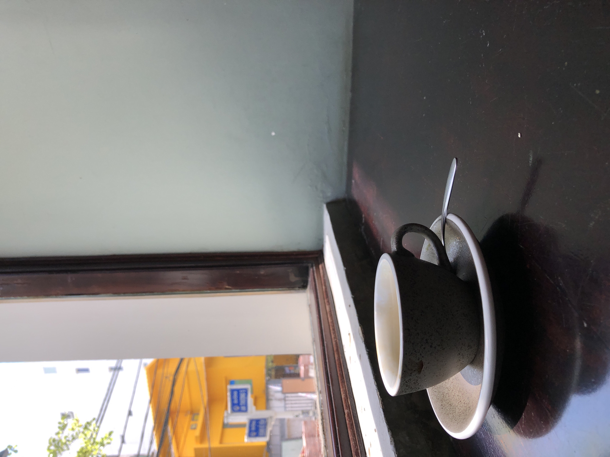 A speckled gray ceramic cup filled with coffee rests on a matching saucer. A silver spoon is positioned on the saucer, partially extending from beneath the cup. The cup and saucer are placed on a dark, reflective wooden surface. Behind the cup, a window frame with dark brown trim is visible, revealing a bright outdoor scene. Through the window, a yellow building with signage is visible, along with trees and a white building. The lighting suggests it is daytime. The image appears to be taken from inside a cafe in Da Nang, Vietnam, given the architectural and urban elements visible through the window.