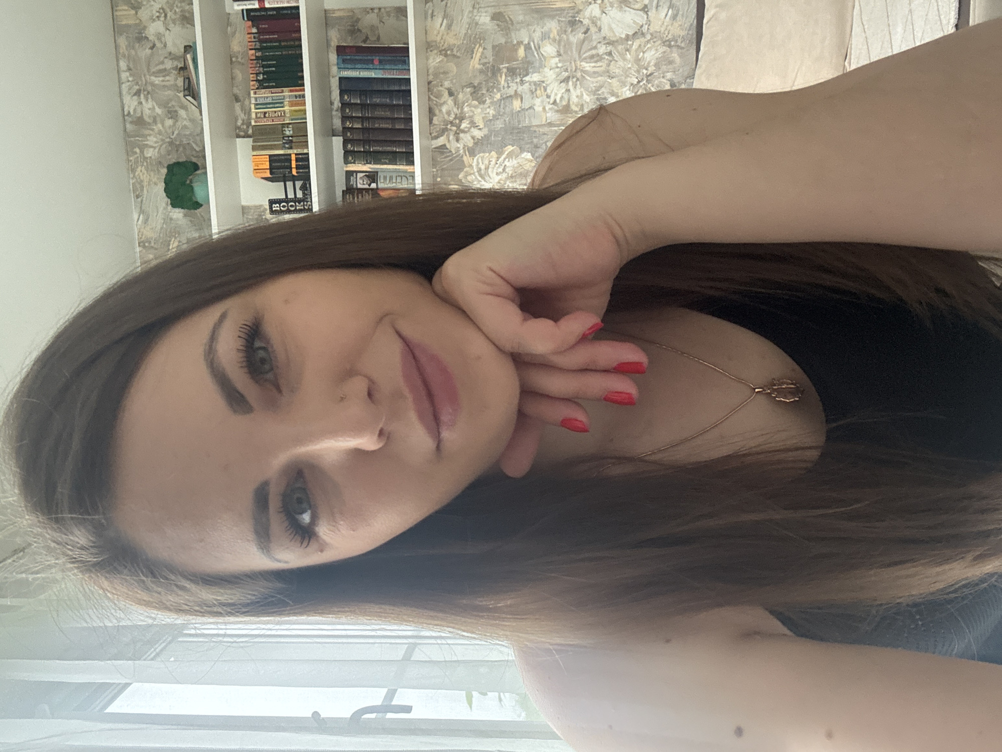 A woman with long brown hair and red nail polish poses indoors. Her left hand rests on her face. She wears a black top and a gold necklace. Behind her is a bookshelf filled with books and wallpaper with a floral pattern. A window is visible in the lower portion of the image. There is not enough information to verify that the photo was taken in Раздолье, Russia.
