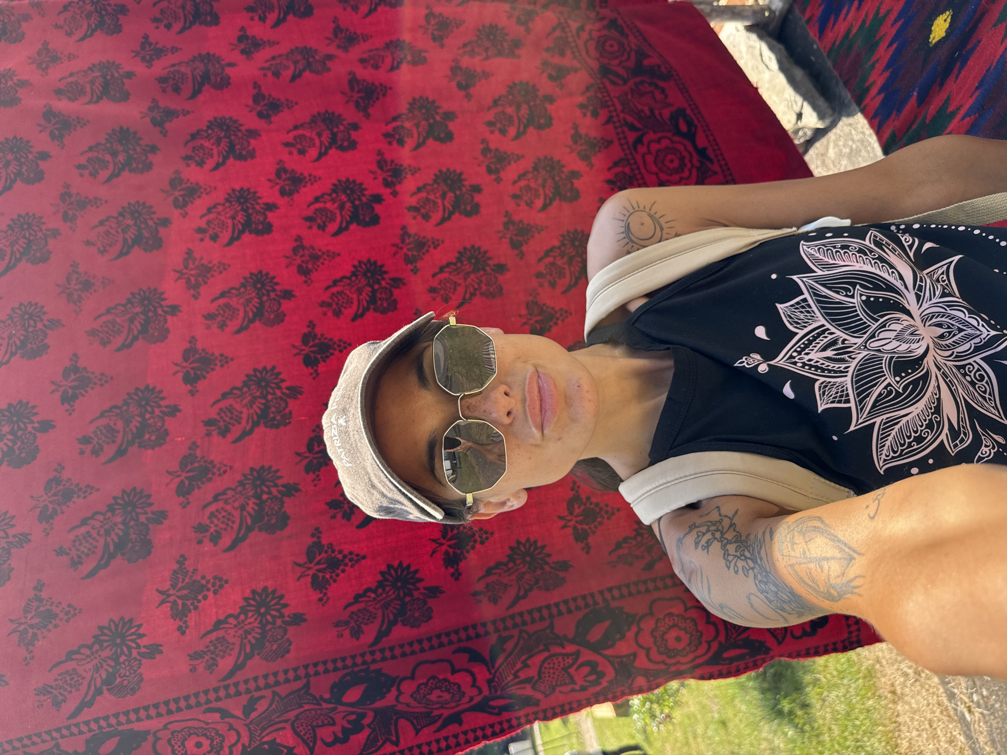 A woman in a baseball cap, sunglasses, and a black tank top with a lotus flower design takes a selfie outdoors. She has tattoos on her arm, including a crescent moon and a sun design. A beige backpack is slung over her shoulder. Behind her is a red fabric with a black floral pattern, and to her right is a colorful rug. The scene appears to be in Ribaritsa, Bulgaria, given the presence of traditional Bulgarian folk art motifs.