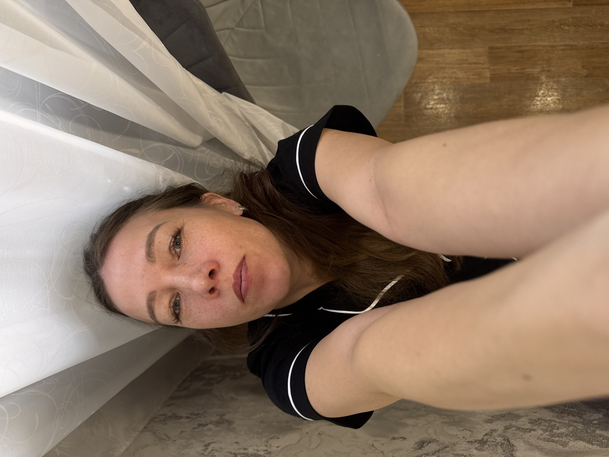 A woman is lying on a floor indoors next to a curtain. The floor is a light brown wood laminate. The woman has fair skin, brown hair, and is wearing a black top with white trim. The location is implied to be in the city of Chaikovsky, Russia.
