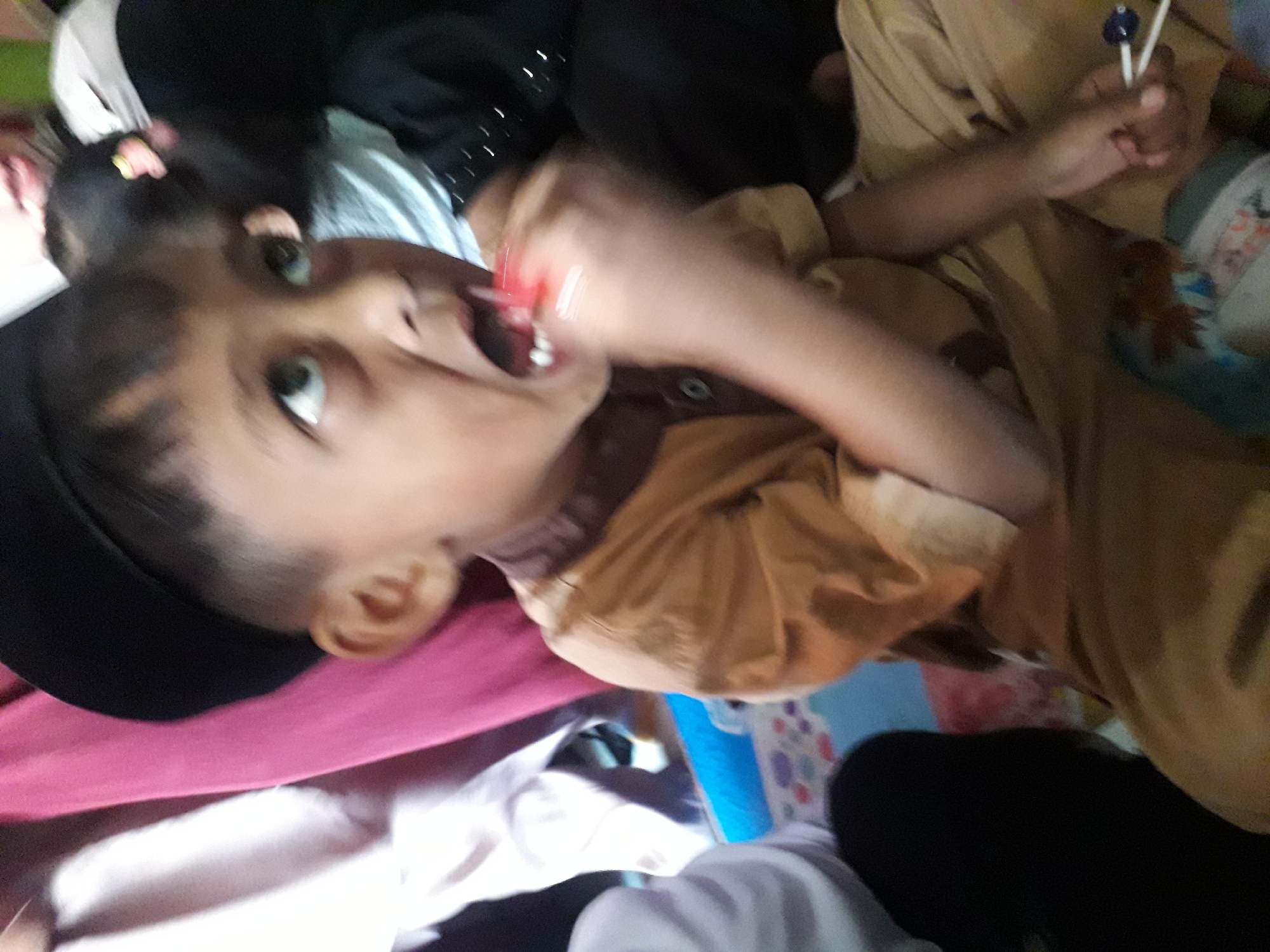A young boy wearing a black skullcap and a tan shirt is in the foreground, holding a red lollipop to his mouth. His eyes are looking upwards and to the right. He is sitting on a patterned mat. In the background, other people are partially visible, including a woman in a pink garment and another person in a black garment. The setting appears to be an indoor gathering, possibly in a home, in Cangkang, Indonesia.
