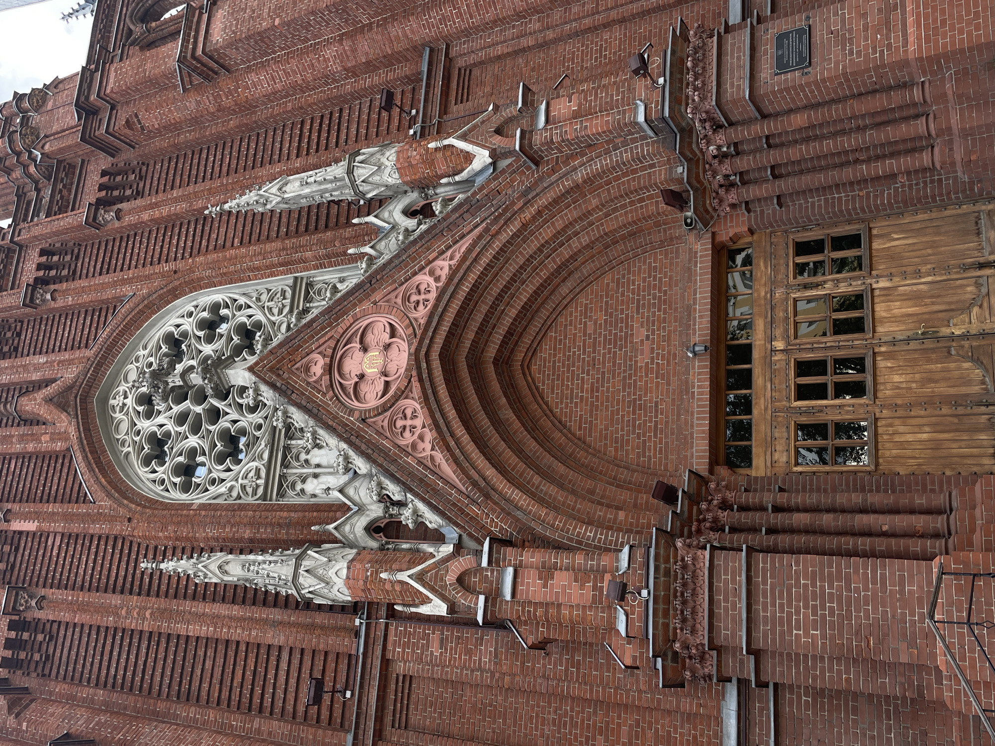 The image shows the entrance of the Roman Catholic Cathedral of the Immaculate Conception in Moscow, Russia. The facade is constructed of red brick and features Gothic architectural elements. A large rose window with intricate stone tracery is visible above a deeply recessed arched doorway. The doorway is framed by ornate brickwork and flanked by pointed spires. A large wooden double door with multiple glass panes forms the entrance. Several exterior lights are mounted on the building. The overall impression is one of grandeur and historical significance.