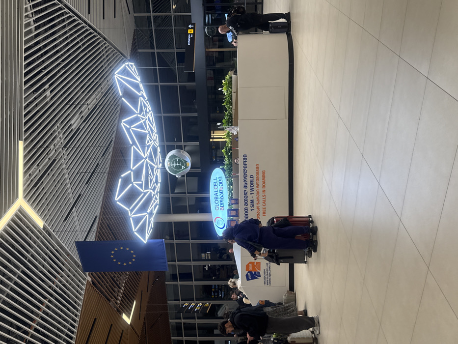 The image shows an airport setting with modern architectural elements. A large, illuminated geometric installation hangs above, near an EU flag. A Globalcell information booth is visible, adorned with plants. Text on the booth promotes “1 SIM - 1 WORLD” and “Free Calls in Roaming.” People with luggage are interacting near the booth. The overall impression is that this is an international arrival or departure area.