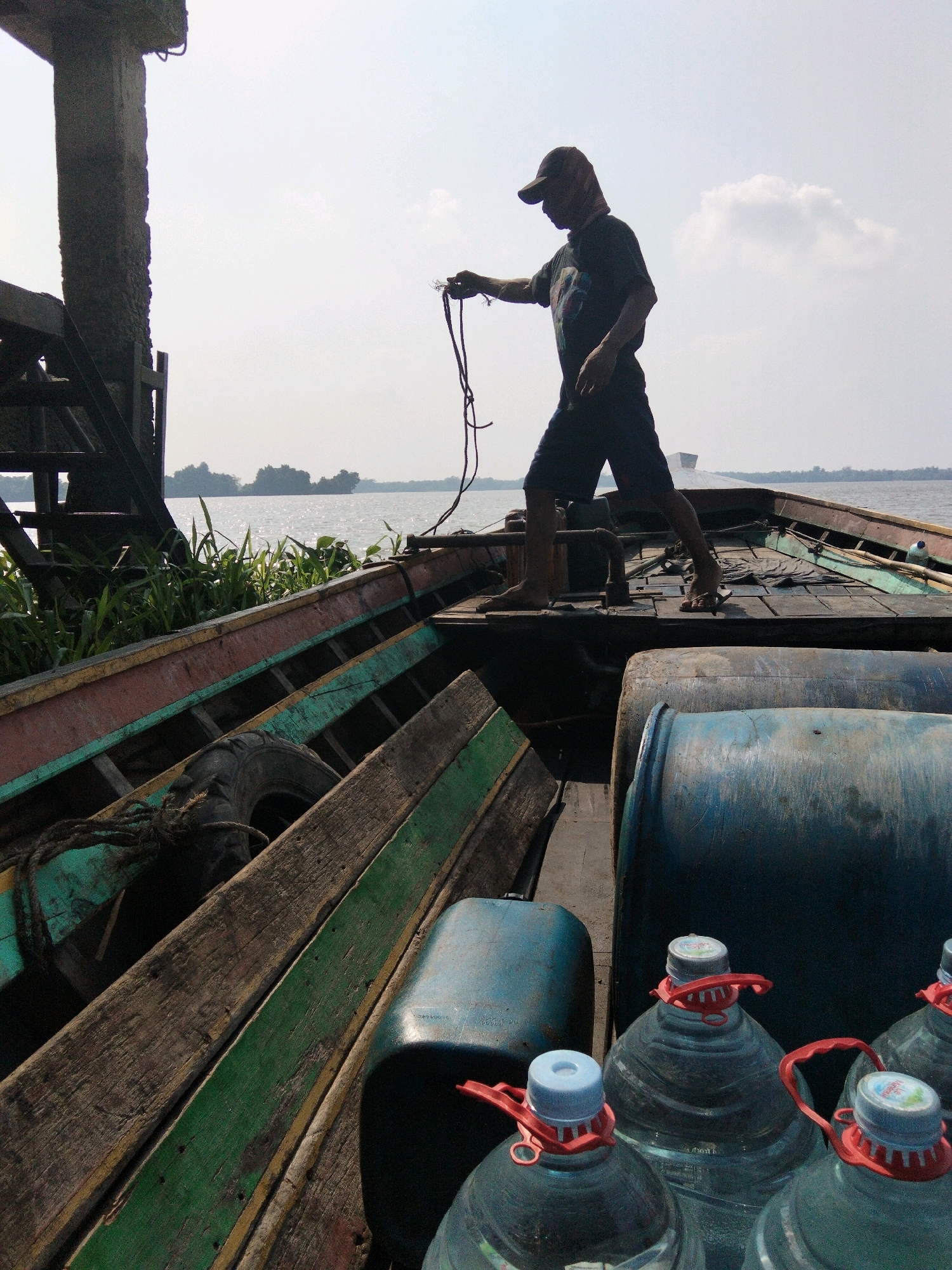 A man wearing a cap and dark t-shirt and shorts stands on a wooden boat loaded with large blue plastic containers, some filled with water. He holds a rope in his hands. The boat is on a body of water with vegetation on the shore and trees in the distance. The scene appears to be in a rural area, likely Indonesia, given the presence of the boat and the natural environment. The sky is bright and cloudy.