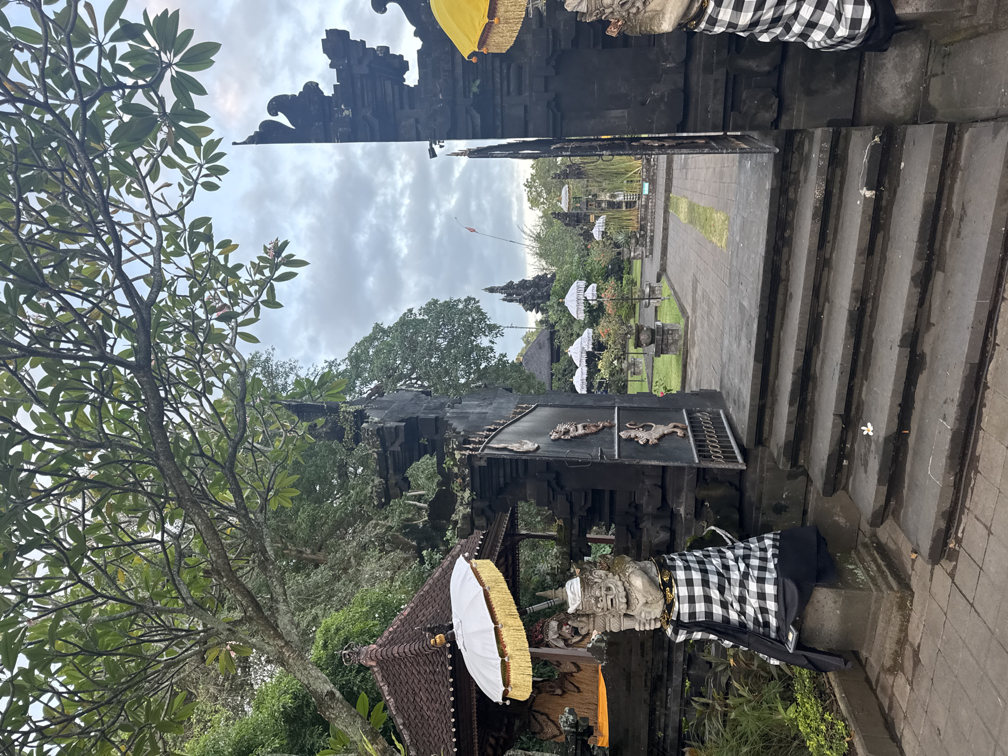 The image shows an outdoor scene at a temple entrance, likely in Dawan, Indonesia. In the foreground, a large stone gate with intricate carvings is partially open, revealing a courtyard beyond. Two stone statues, dressed in traditional black and white checkered cloth, flank the entrance. One statue holds a white umbrella with gold trim. Behind the statues, a traditional Balinese structure with a dark tiled roof is visible, featuring a white umbrella and ornate carvings. The courtyard itself contains several more white umbrellas arranged in a garden setting, with lush green trees and vegetation surrounding the area. In the distance, more temple structures and a flagpole can be seen against a cloudy sky. The ground is paved with stone tiles and there are stone steps leading up to the gate. A single white flower lies on one of the steps.