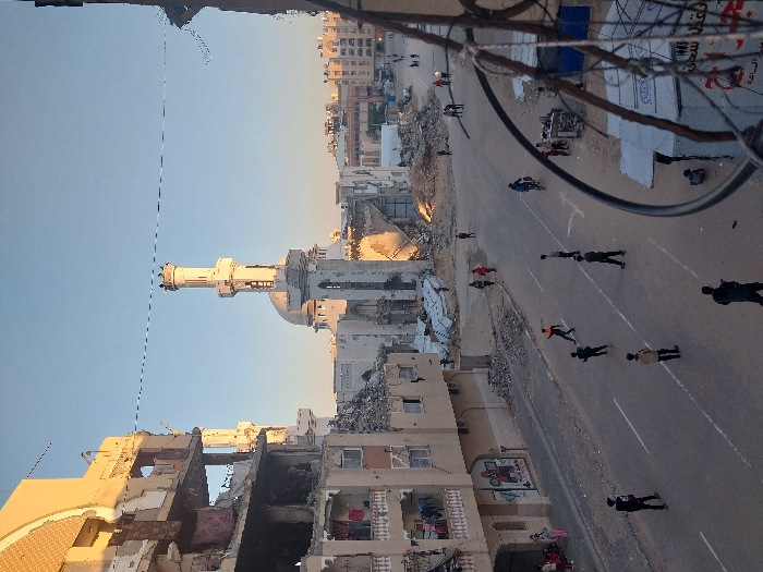 The image displays an urban street scene in Khan Yunis, Palestinian Territory, characterized by widespread structural damage under a clear, bright sky. The prominent central feature is the partially destroyed Al-Farouq Mosque, identified by its main dome and two minarets, one of which stands largely intact while the other shows damage. Substantial piles of rubble, concrete, and twisted metal are concentrated around the mosque and adjacent areas, indicating significant collapse.
Along both sides of the paved street, which retains its white lane markings, multiple multi-story buildings exhibit severe damage, including exposed interiors, partial floor collapses, and visible rebar. Laundry is visible hanging from a balcony of a damaged building on the left.
Numerous individuals, primarily appearing as young males or children, are present on the street. Observed actions include people walking, standing in small groups, and one individual riding a bicycle towards the right. One person wearing an orange top is depicted in a dynamic pose, with arms raised, on the street. In the immediate foreground on the right, utility wires and a section of a makeshift or temporary shelter are visible. The lighting suggests either early morning or late afternoon, casting soft shadows.