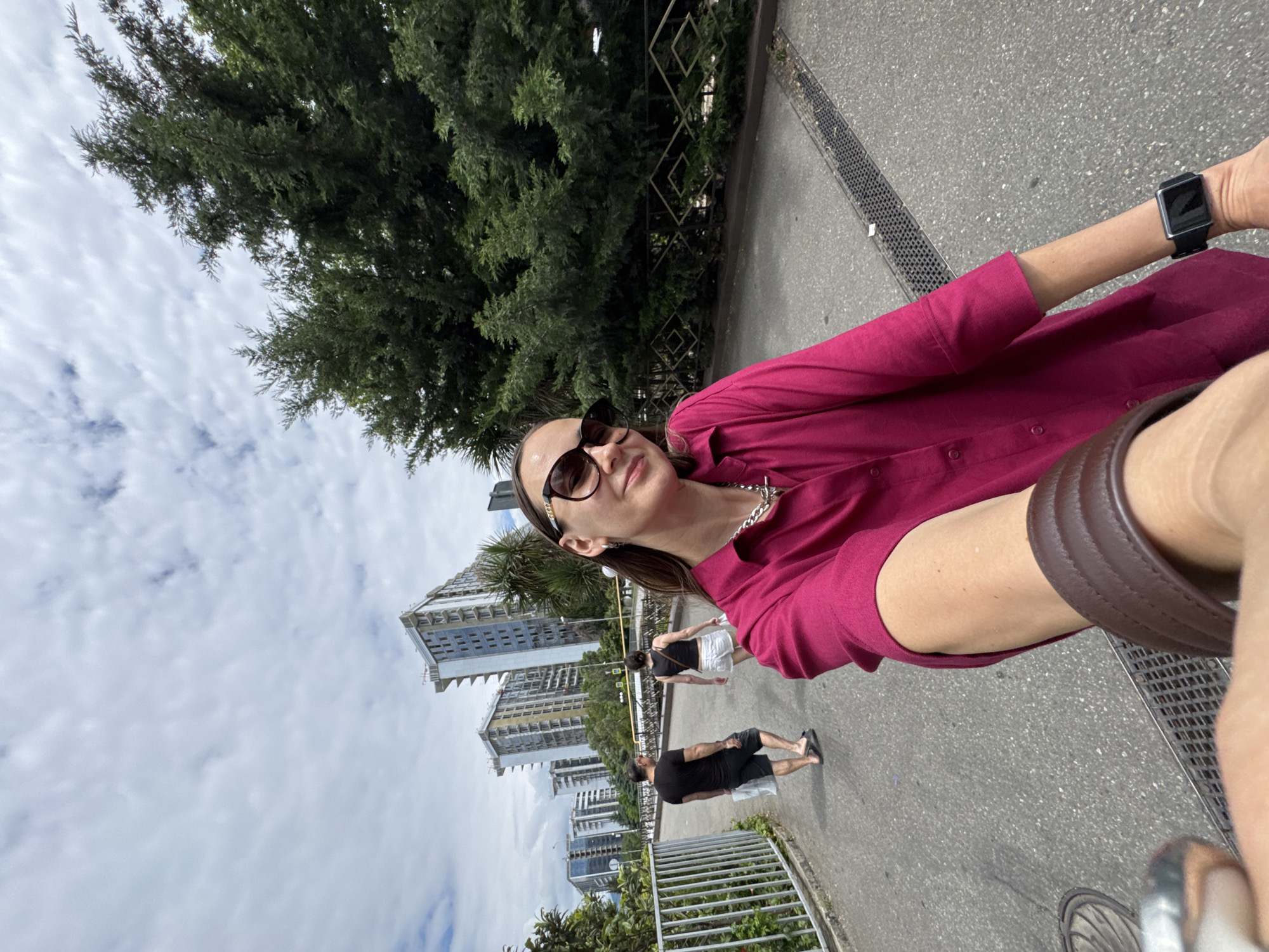 An outdoor, wide-angle, first-person perspective selfie shows a woman with sunglasses and a pink collared shirt walking on a paved path in Sochi, Russia. Behind her, a man in a black t-shirt and shorts walks away from the camera, holding two white bags. A woman in a black tank top and white shorts is walking in the same direction as the man. In the background, several modern, tall buildings under construction are visible. Lush green trees flank the path on the right. The sky is cloudy and overcast. The woman in the foreground is wearing a silver necklace and a smart watch on her left wrist.