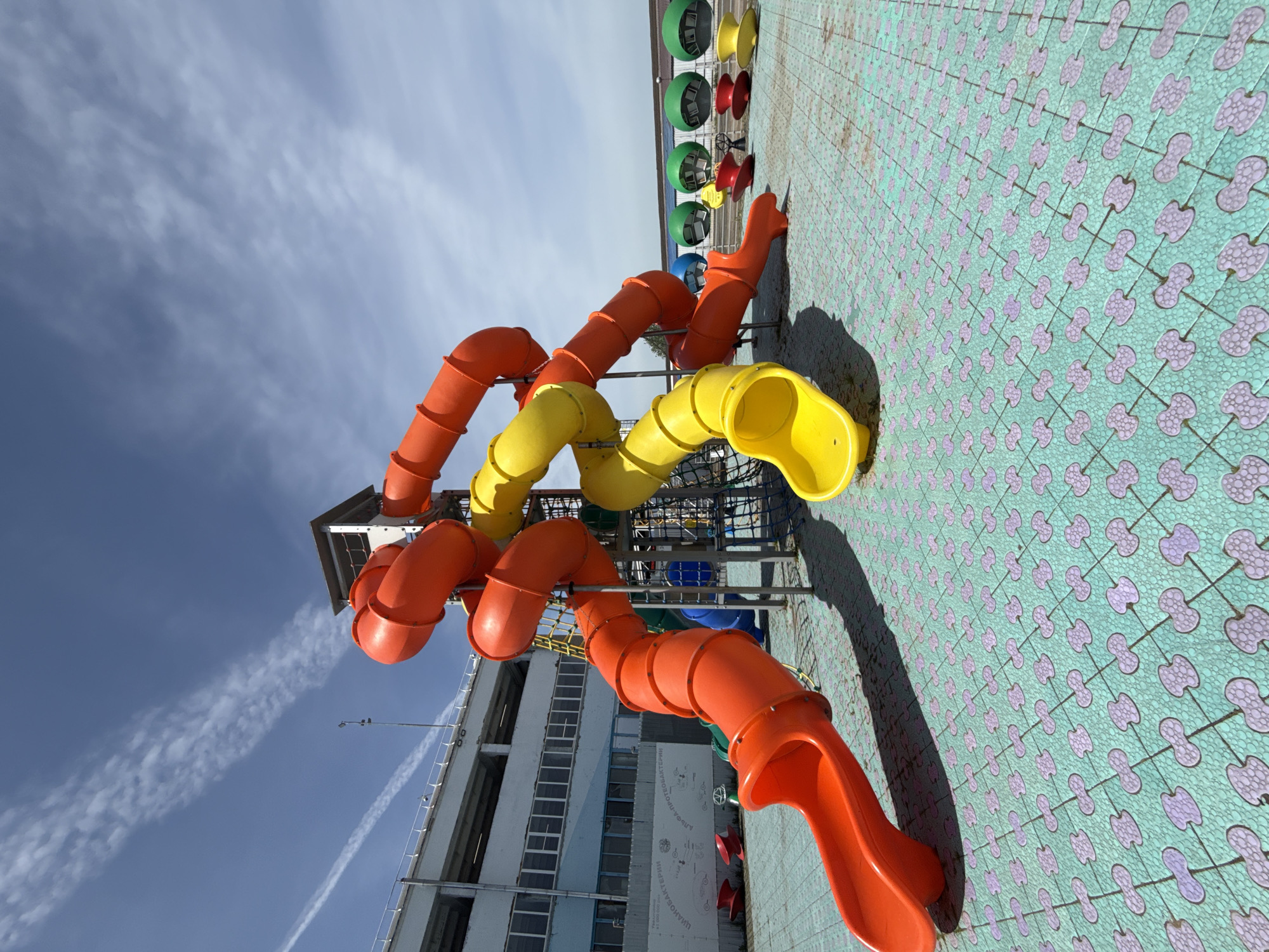 The image shows an outdoor playground in Синьково, Russia, featuring colorful slides - orange and yellow - connected to a structure against a backdrop of a blue sky with clouds. The ground is covered in patterned tiles. A building is visible in the background.