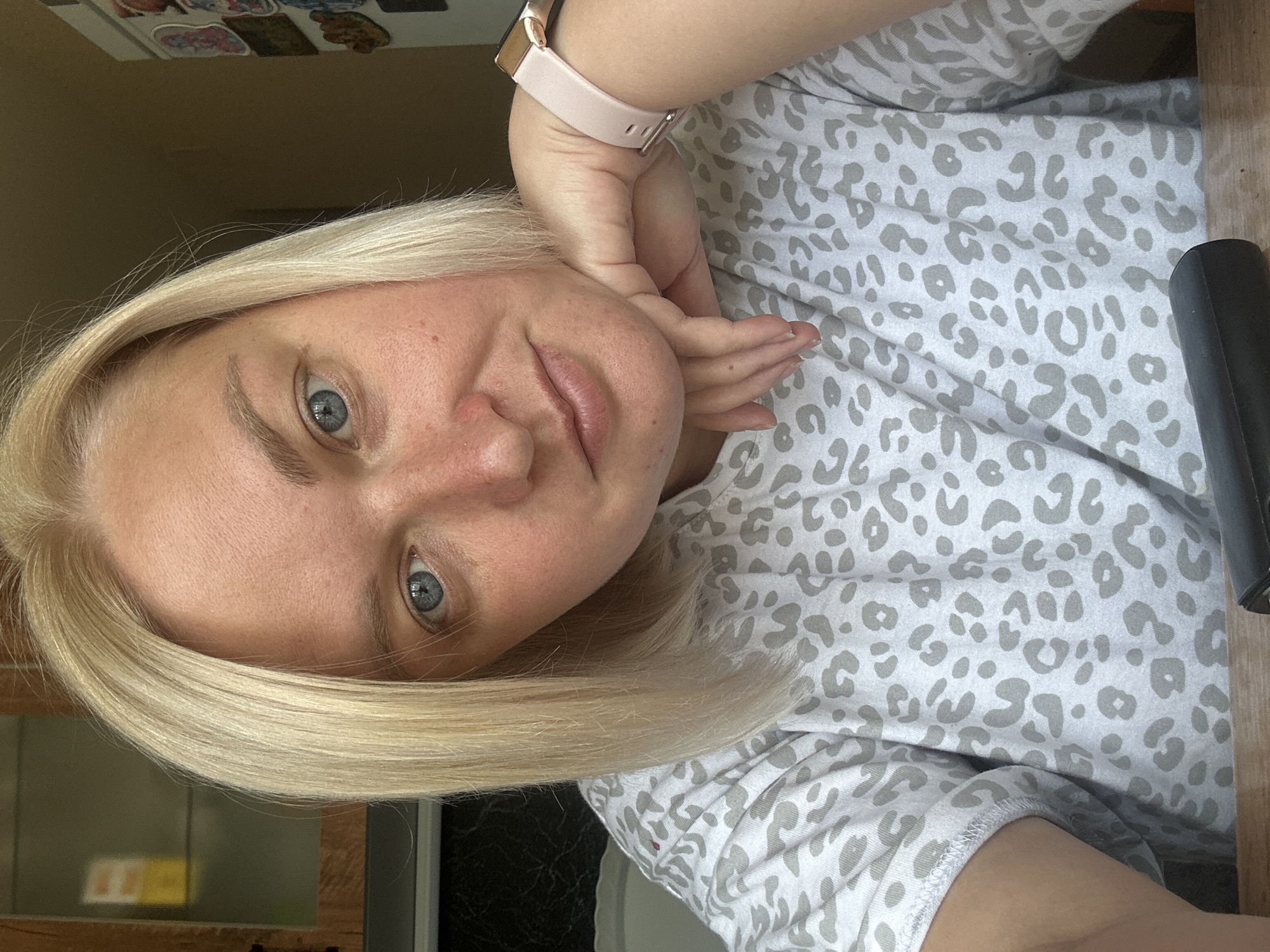 A person is shown in a close-up, angled selfie. They have blonde hair and blue eyes, and are wearing a grey t-shirt with a leopard print pattern. Their right arm is bent, and their hand is resting on their cheek, with a light pink smartwatch on their wrist. The background is blurred, but appears to be an indoor setting with wooden furniture visible on the right. The lighting suggests it could be daytime. The person's expression is neutral or slightly smiling. There is no visible text in the image. The metadata indicates the location is Chaikovsky, Russia.