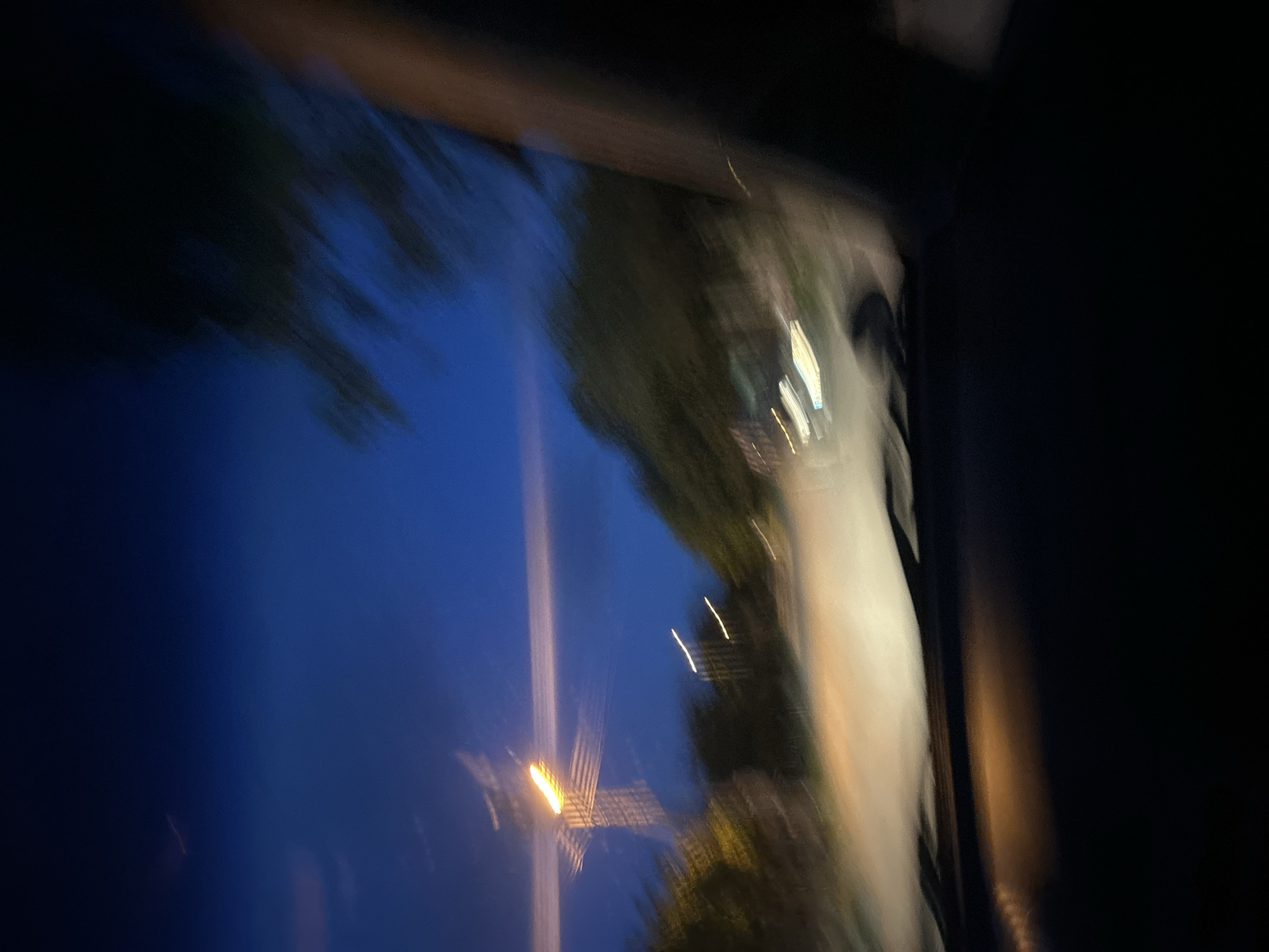 The image shows a blurred view from inside a vehicle at night or dusk. The left side of the frame features a dark blue sky with blurred streetlights. On the right side, a light-colored road is visible, bordered by trees or foliage. The image is out of focus and displays motion blur. The location, Pishtigovo, Bulgaria, cannot be confirmed from the visual data.