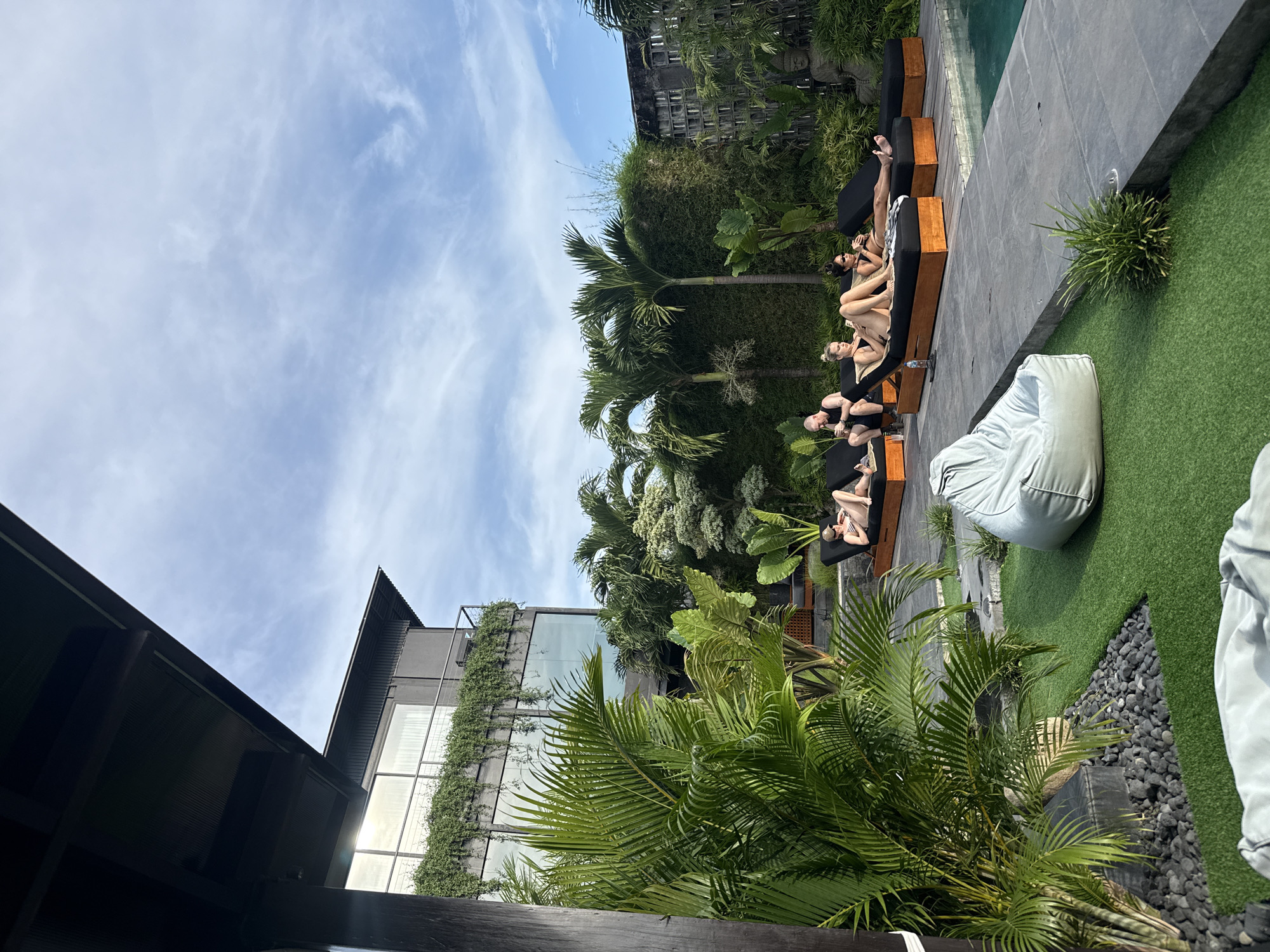 The image captures a vibrant, sunny outdoor scene at what appears to be a resort or luxury residence. The primary subjects are several individuals, mostly women, dressed in swimwear, lounging on dark, cushioned daybeds next to a swimming pool. They are positioned along the edge of the pool, seemingly relaxing or sunbathing.
The setting is characterized by lush tropical landscaping, with numerous palm trees and other dense green foliage dominating the midground and background. Artificial turf covers an area adjacent to the pool, where a large, light grey bean bag chair is situated. Dark grey paving stones border the pool, and an area of dark pebbles separates the paved section from the green turf.
In the background, a modern multi-story building with dark facades and large windows is visible, partially adorned with climbing green vines. Another building or structure, partially obscured by trees, features what looks like a stone statue, possibly of a Buddha head, adding to the serene atmosphere.
The time of day is bright daytime, indicated by strong sunlight and clear blue skies with wispy white clouds. The overall mood is one of leisure and tranquility. No visible text is present in the image.