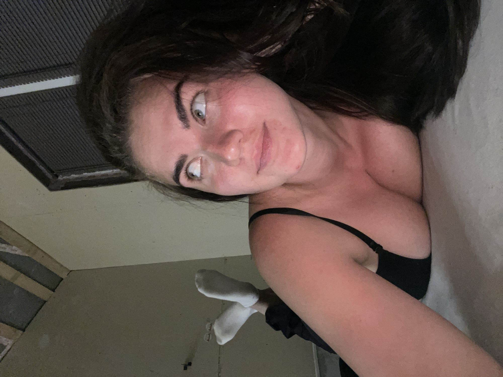 The image shows a woman lying on a bed, looking up and to the right. She has long, dark hair and is wearing a black camisole and white socks. The room appears to be unfinished, with exposed ceiling beams and unpainted walls. There is a window with blinds in the background. The overall lighting is dim. The specific location in Мосметаллоконструкция Онт, Russia, cannot be determined from the image alone.