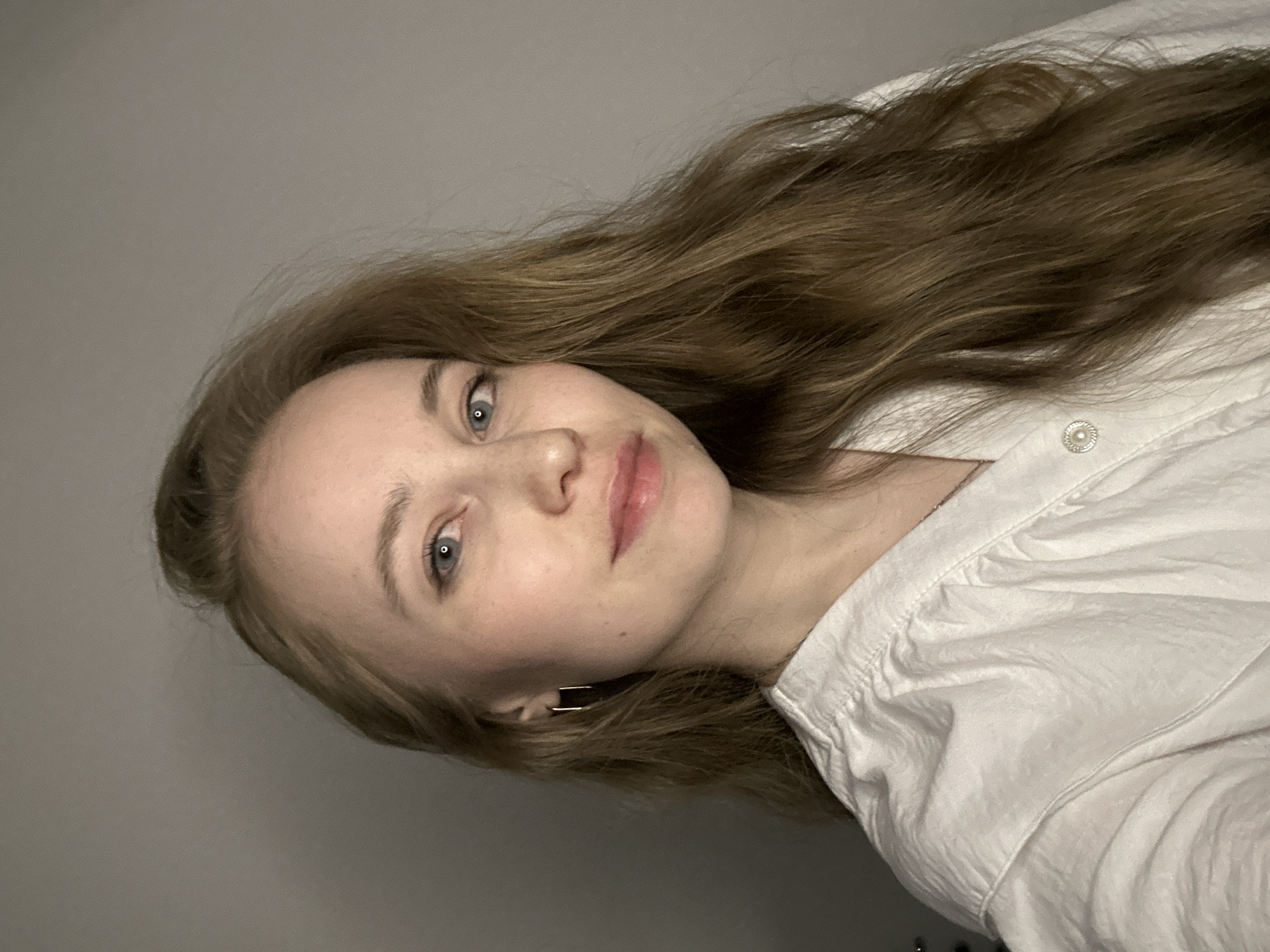 The image features a rotated portrait of a young woman, likely in her late teens or early twenties, against a plain, light gray background. Her head is positioned to the left, and her body extends to the right, implying a 90-degree counter-clockwise rotation of the photograph.
The woman has long, wavy, light brown hair with subtle reddish highlights, which is spread out around her head. She possesses fair skin, light blue eyes, and subtle eyebrows. Her lips are slightly parted, revealing a soft, natural pink color, and she has a gentle, calm expression with a hint of a smile.
She is dressed in a white collared or button-down shirt, which appears to be made of a lightweight fabric like cotton or linen and shows some natural folds. A small, round, pearl-like button or decorative embellishment is visible on the upper part of her shirt, near the shoulder. A small, rectangular, gold-toned earring is visible in her left ear.
The setting is indoors, with a minimalistic, plain background that provides no specific environmental details. The lighting is soft and even, suggesting either natural light from an unseen window or diffused artificial lighting. The woman is posing for the camera. There are no visible location cues within the image itself that would identify it as Saint Petersburg, Russia, nor is any text present. The overall impression is one of peacefulness and simplicity.