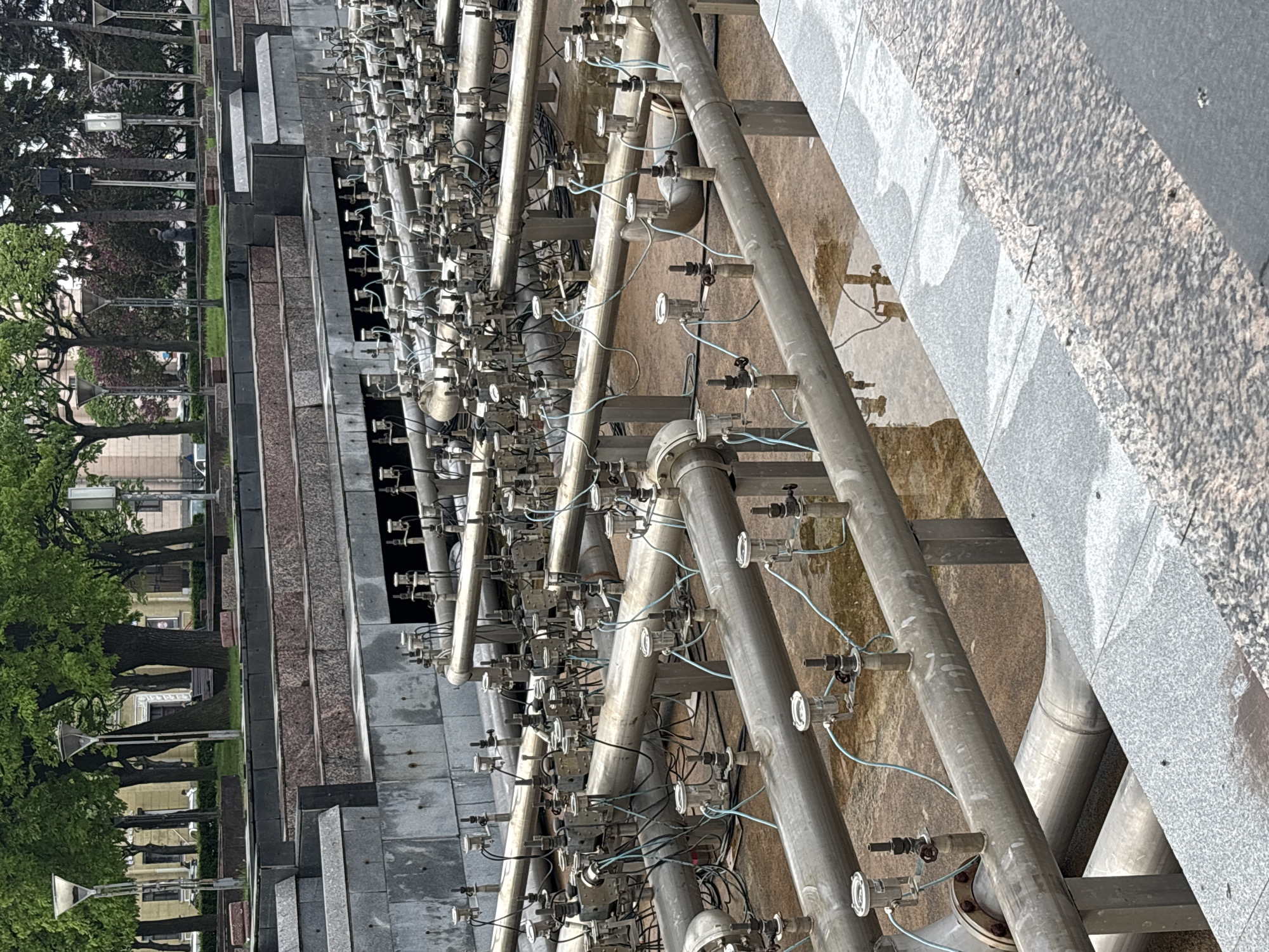 The image captures a section of the Saint Petersburg Fountain Square fountain system. The fountain features a complex network of metal pipes with numerous nozzles, valves, and electrical connections. The base of the fountain is constructed from gray and pink granite blocks arranged in a stepped design. In the background, there are trees, green grass, and street lamps indicating a public park area. There are no people visible in the immediate vicinity of the fountain. The overall environment suggests an urban public space.