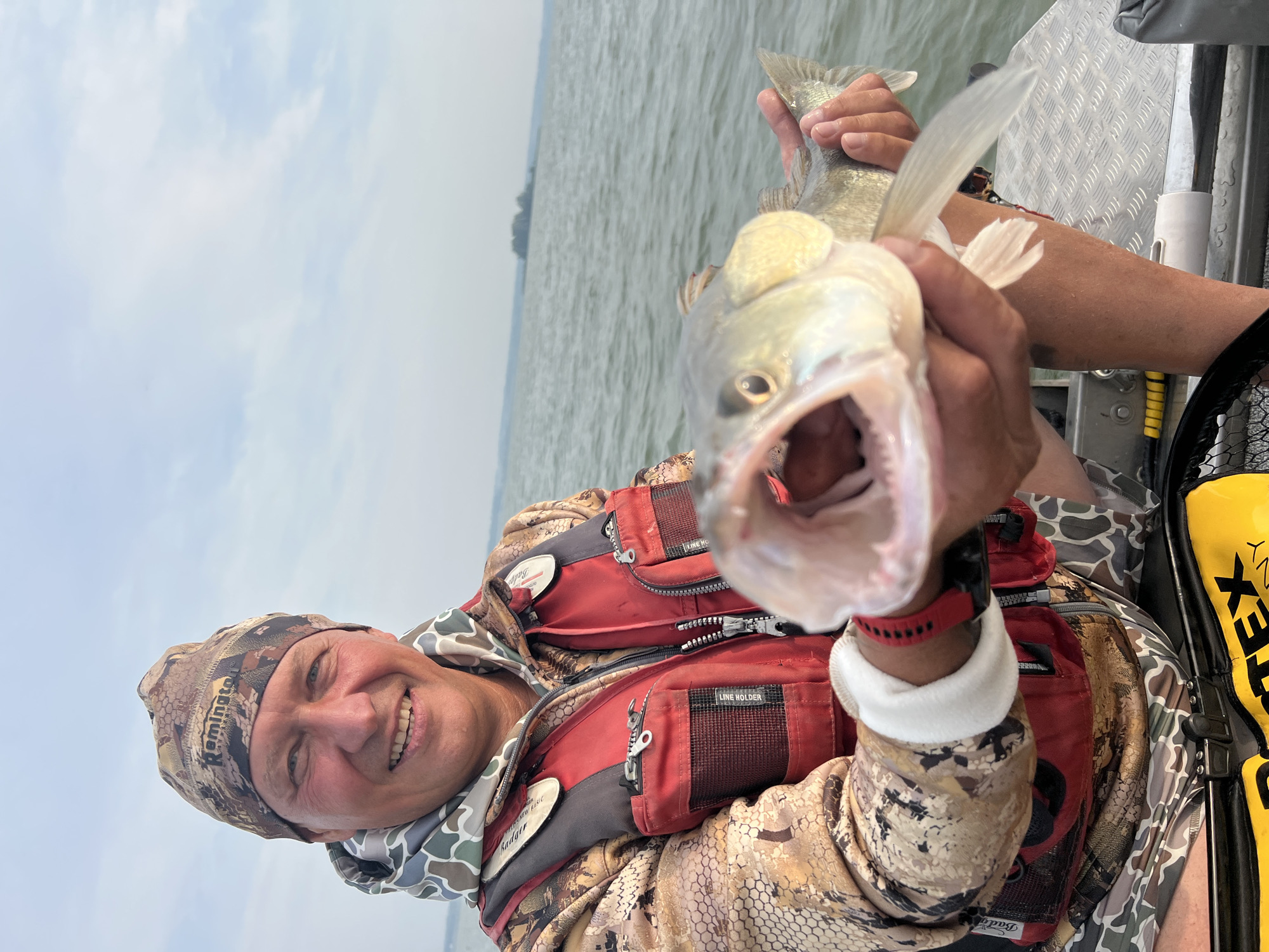 A man in camouflage clothing and a red life vest holds a large fish in front of him on a boat. The man is smiling at the camera, and the fish's mouth is wide open, revealing its teeth. The boat appears to be on a large body of water with a distant island visible on the horizon. The sky is overcast with scattered clouds. The man is wearing a camouflage beanie with the "Remington" logo visible. The life vest has multiple pockets and zippers. The boat's deck is made of diamond-plate metal. In the foreground, a yellow bag with the word "Vortex" is partially visible. The setting is likely the Ob River near Novopichugovo, Russia, a region known for its fishing opportunities.