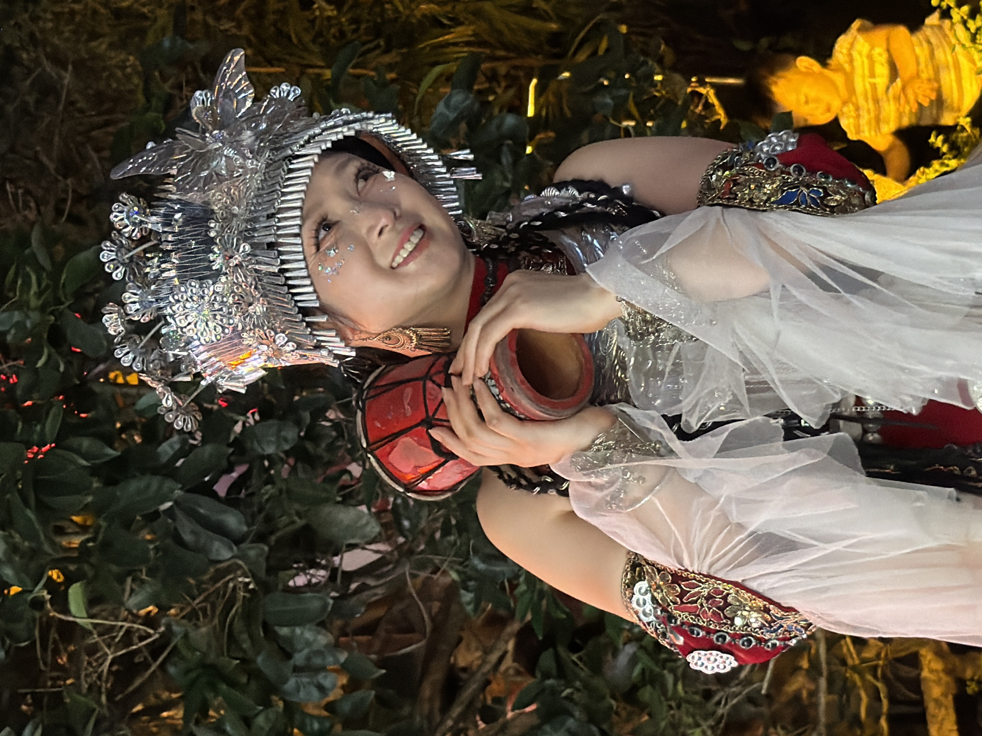 In this image, a woman in elaborate traditional attire is the central subject. She wears a large, ornate headdress adorned with silver, crystalline elements, and sparkling facial makeup. Her costume includes a red, sequined, and jeweled band around her arm, and she is holding a small, red drum in her hands. The scene appears to be set outdoors, with lush green foliage forming a backdrop. In the upper right portion of the image, a blurred, yellowish light suggests artificial lighting or perhaps a distant illuminated structure. The overall lighting suggests it is likely evening or nighttime, adding a dramatic ambiance. The woman's expression is one of joy, with a bright smile and eyes looking upwards. The location context is Yuzhong District, China, though no specific landmarks are identifiable in the image itself. The media depicts a moment of performance or celebration, possibly a cultural event.