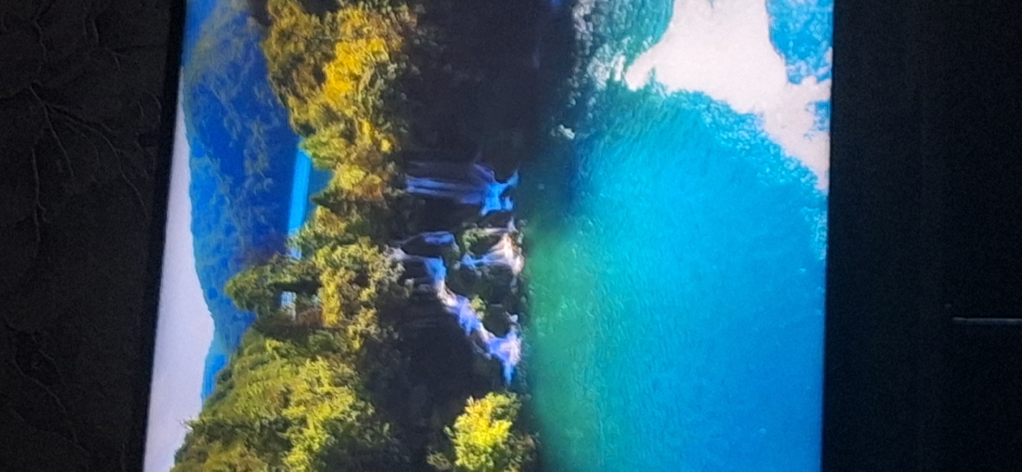 The image depicts a serene natural landscape featuring lush green trees, cascading waterfalls, and a clear turquoise body of water. The background shows rolling hills covered in blue foliage. There are no people or man-made structures visible in the scene. The location is identified as being in the city of Bila Tserkva, Ukraine.
