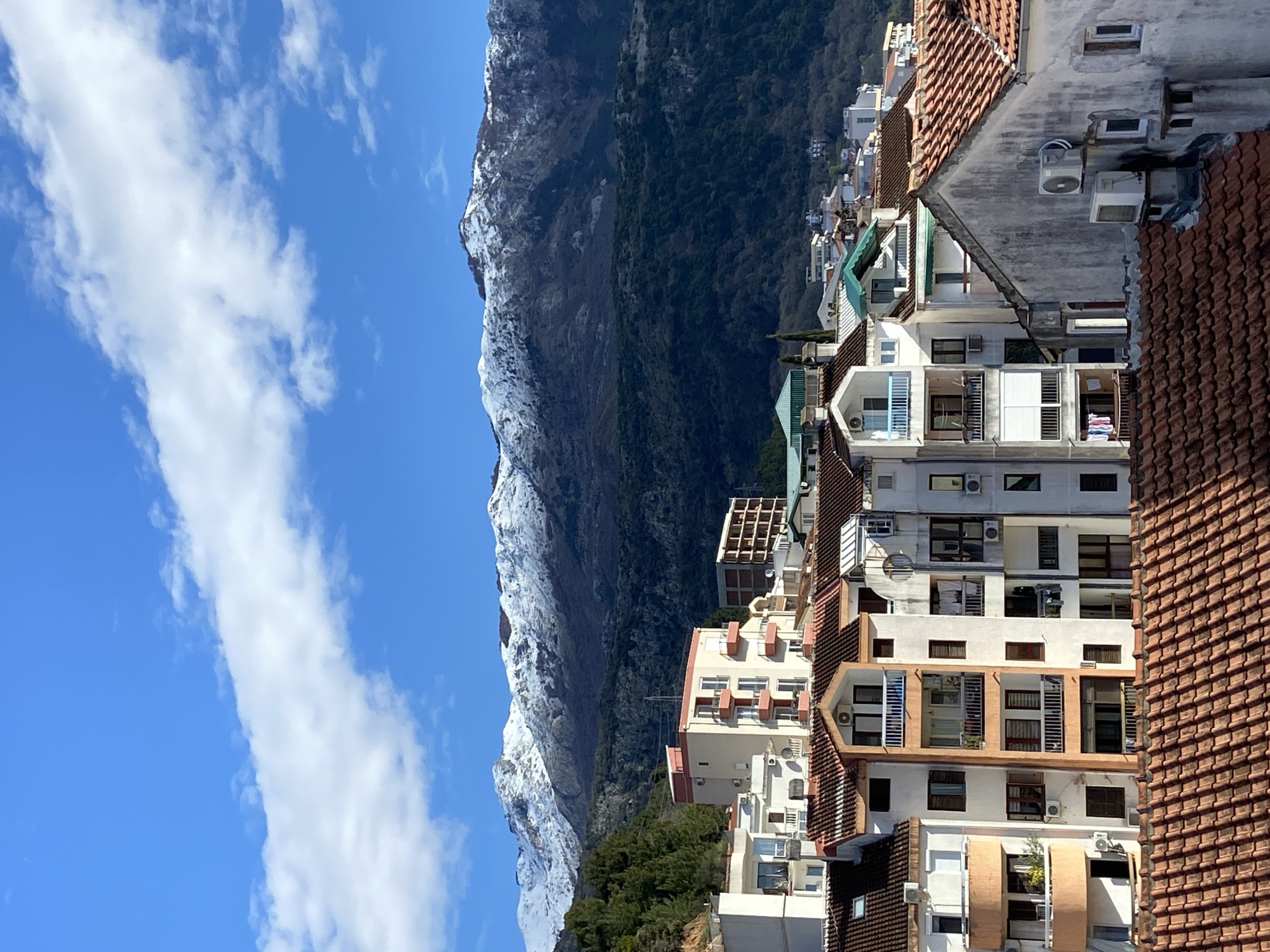 The image depicts a mountain range partially covered with snow, viewed from the city of Budva, Montenegro. The sky is blue with scattered white clouds. In the foreground, there are apartment buildings with a variety of architectural styles and colors, featuring tile roofs, balconies, and air conditioning units. The mountains serve as a backdrop to the urban landscape. The scene appears to be taken on a sunny day, showcasing the contrast between the natural environment and the built environment of the coastal city.