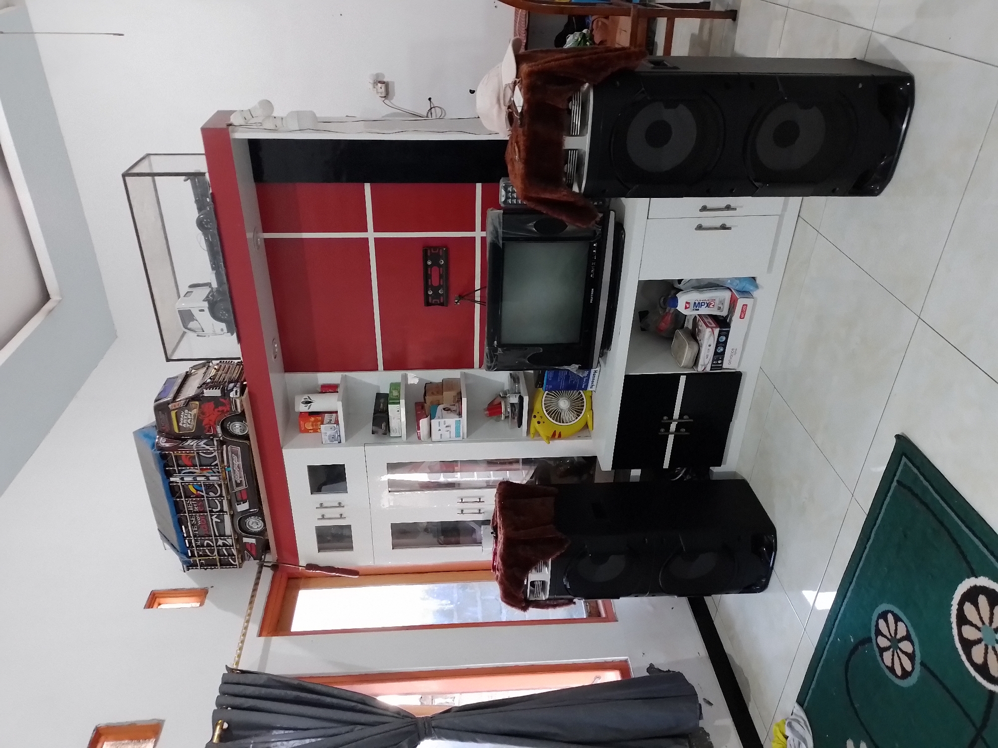 The image shows a room with white walls and tiled flooring, likely in a residential setting in Ngancar, Indonesia. A large entertainment center dominates the right side of the frame, featuring a red and white facade with shelving. On the top shelf, two model trucks are displayed; one is a detailed replica of a colorful Indonesian truck, and the other is a white truck chassis inside a glass case. Below, a television sits on a shelf, flanked by two large black speakers. A yellow oscillating fan is visible on a lower shelf, along with various boxes and containers. To the left of the entertainment center, dark gray curtains hang by a window with wooden framing. The floor is partially covered by a green rug with circular floral patterns. The scene appears to be an indoor living space, possibly a living room or entertainment area.